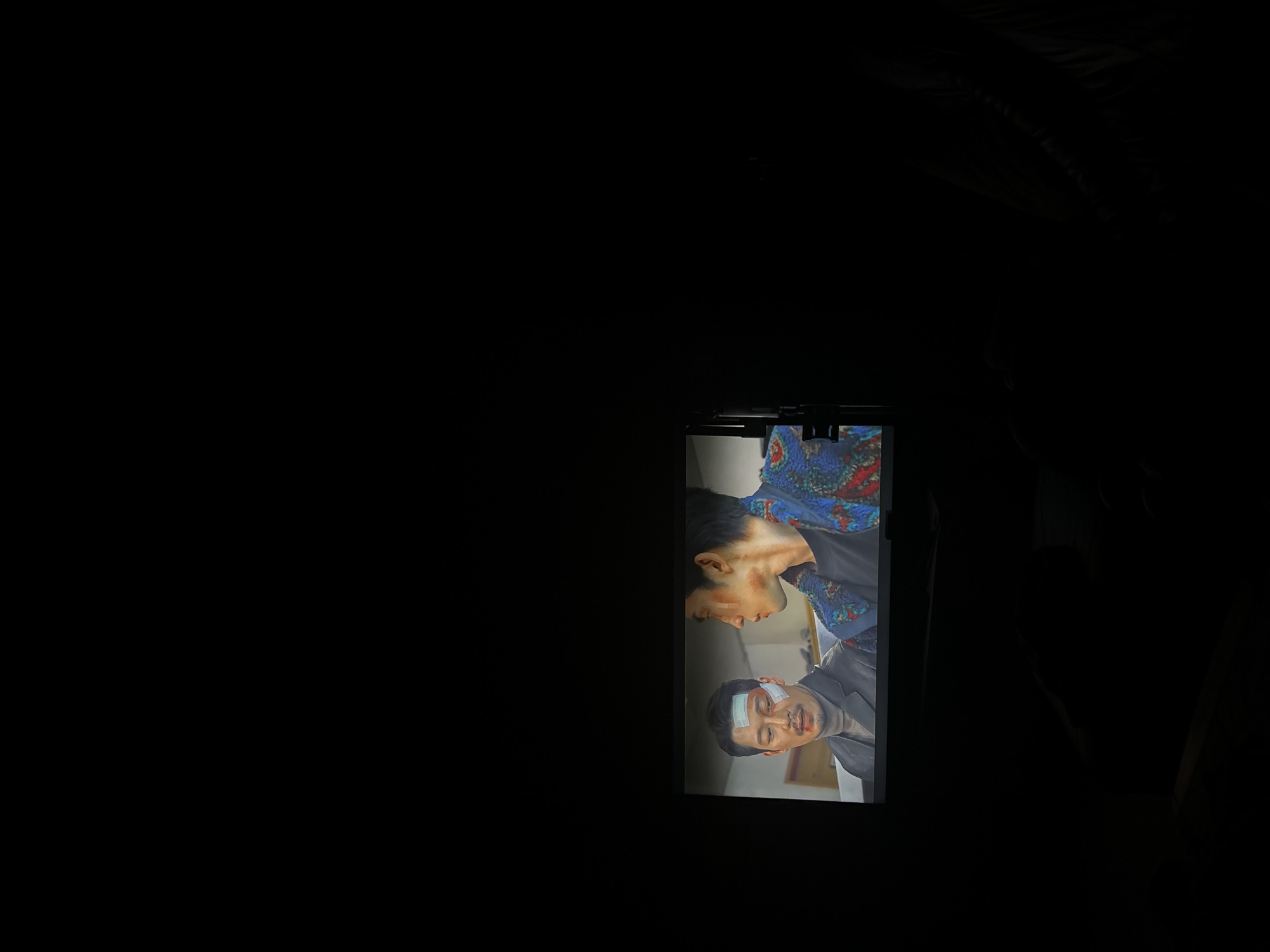A television screen displays a scene from a show or movie, with two men visible. The man on the left has a bandage on his forehead and is wearing a dark turtleneck and blazer. The man on the right is wearing a black t-shirt under a colorful, patterned jacket. The background on the screen is blurred, but appears to be an indoor setting. The television is positioned in a dark room, with the majority of the image being black.