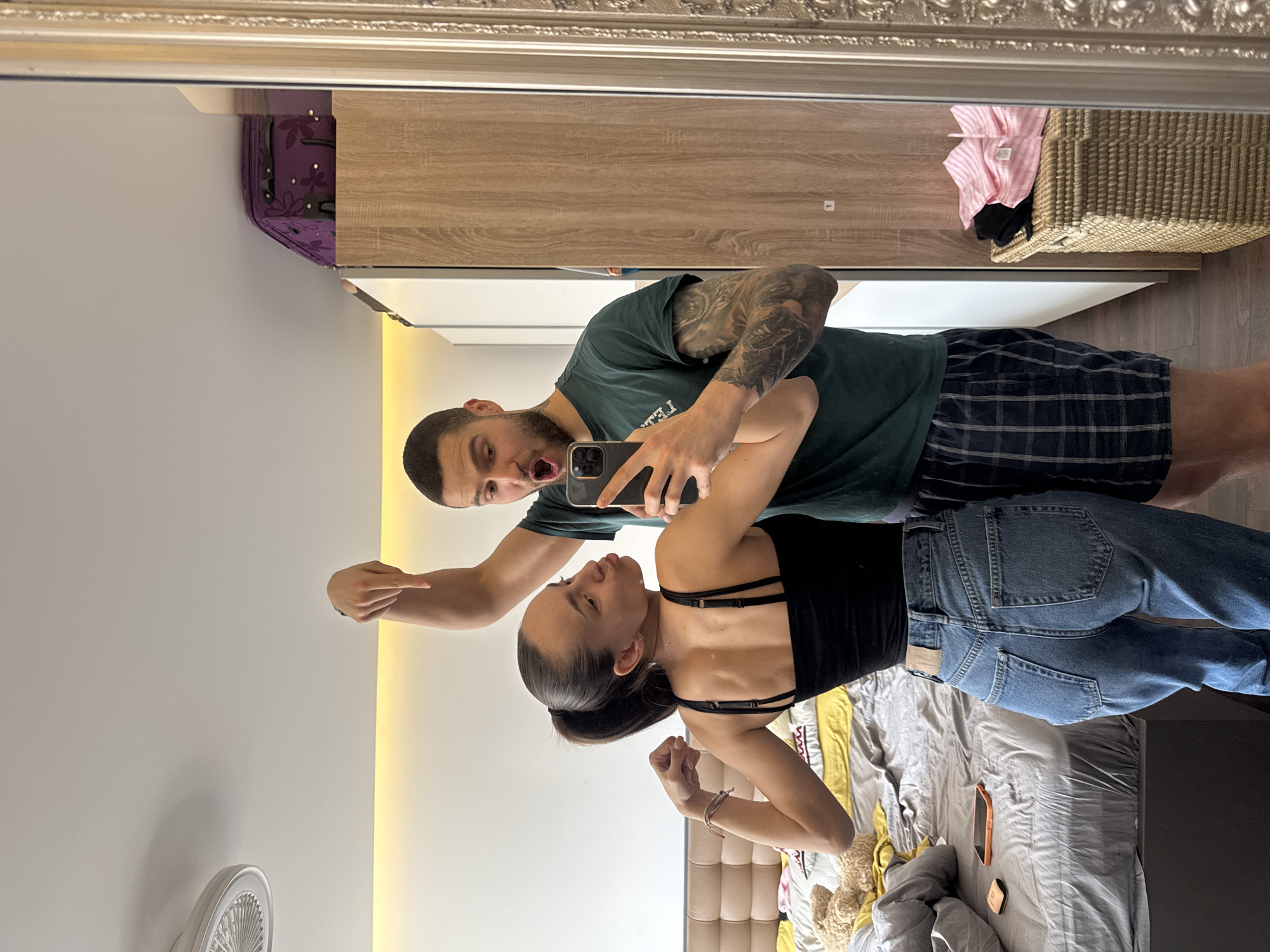 A mirrored image shows a man and a woman. The man, on the left, has dark hair and a beard, and is wearing a dark green t-shirt and dark plaid shorts. He has tattoos on his left arm and is holding a smartphone, presumably taking the photo. His mouth is open in a mock yell, and his left arm is extended, pointing toward the woman. The woman is wearing a black top with thin straps and blue jeans. She has dark hair pulled back into a ponytail. She is posing with her arms flexed. The background includes a light gray wall, a wardrobe with light wood-grain doors, a wicker basket containing clothes, and a bed with rumpled bedding. The top of the mirror's frame is visible. The location is in an interior, probably an apartment or hotel room in Sofia, Bulgaria.