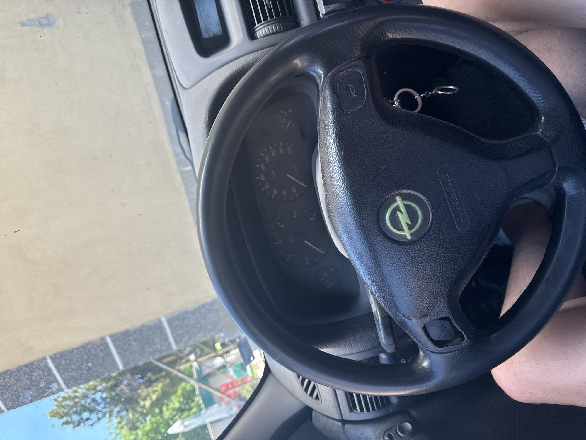 The image shows the interior of a vehicle, specifically the steering wheel. The steering wheel is black and features a green Opel logo in the center. A set of keys hangs from the steering wheel. The dashboard and instrument panel are visible behind the steering wheel. Outside the vehicle, trees and parts of buildings are visible through the windshield, which indicate that the location is Dermantsi, Bulgaria. Parts of a person's leg is seen near the steering wheel.