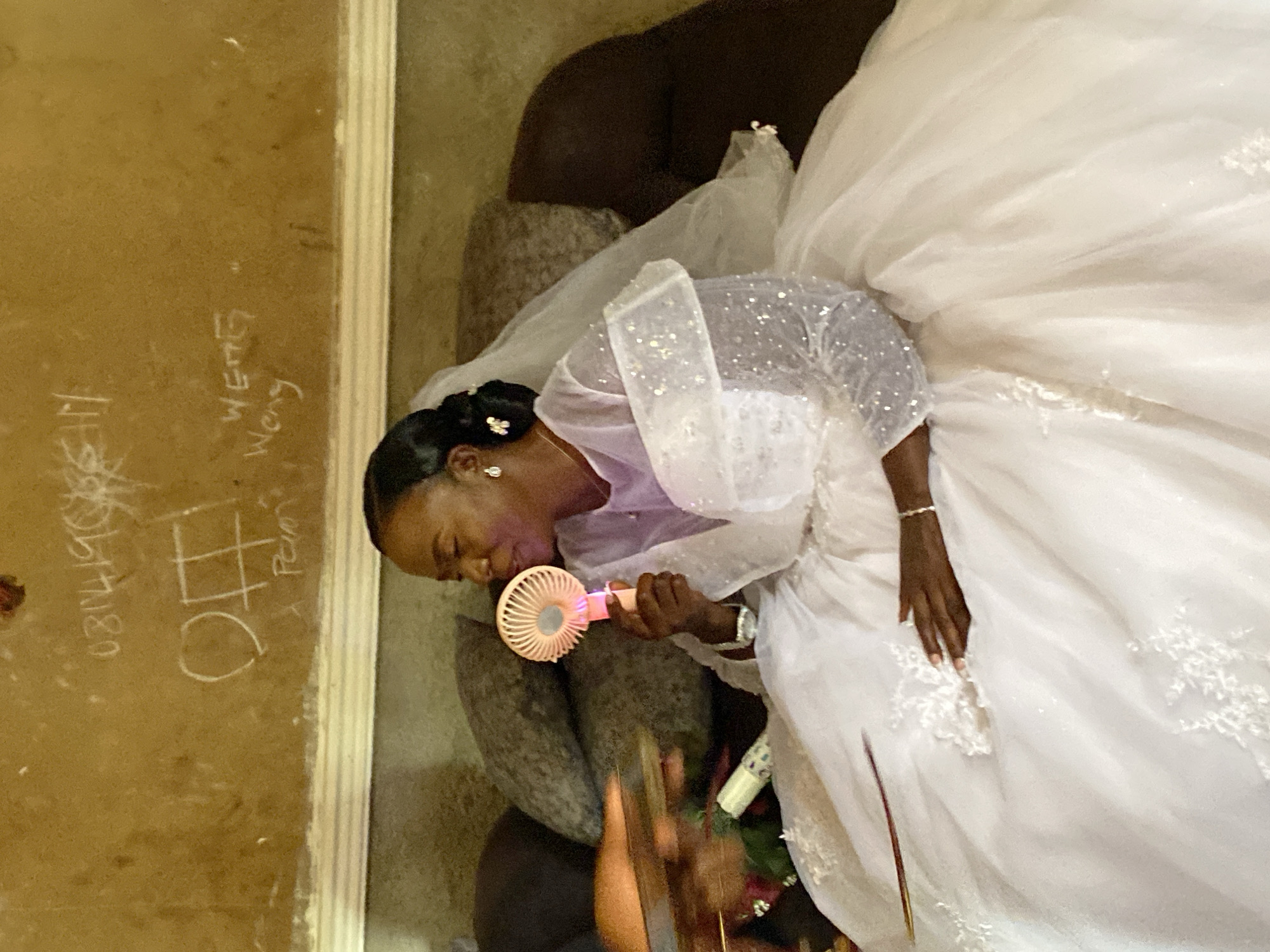A woman in a white wedding dress and sheer, embellished top holds a small, peach-colored fan to her face. She has dark hair pulled back and wears jewelry. The background is a wall with faded graffiti, including numbers and words, and a dark-toned object beside her. The scene appears to be indoors.