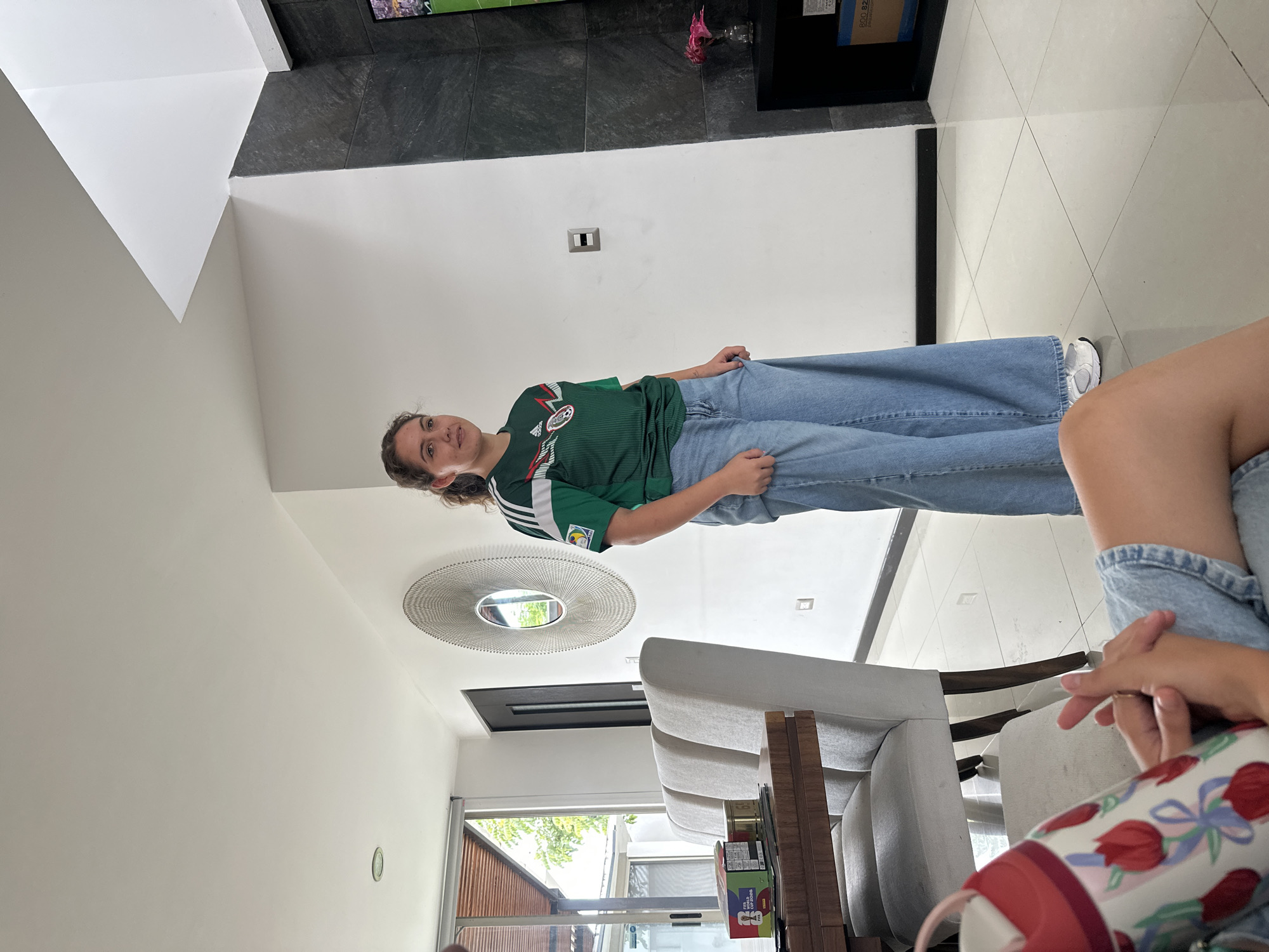 In a well-lit, modern indoor setting, a young woman stands in the center of the frame, wearing a green and white Mexican national soccer team jersey and loose-fitting light blue jeans. Her dark hair is pulled back, and she has a neutral expression. To her left, a wooden dining table with four upholstered chairs is visible, with a few boxes and a tin can on its surface. Behind the woman, a large, circular metallic wall decoration hangs on a white wall. To her right, a dark gray, stone-tiled accent wall rises upwards, flanking a television screen displaying a sports broadcast. The floor is tiled with large, light-colored squares, reflecting the ambient light. The background suggests a residential or hospitality environment within the city of San Bernardino Tlaxcalancingo, Mexico.