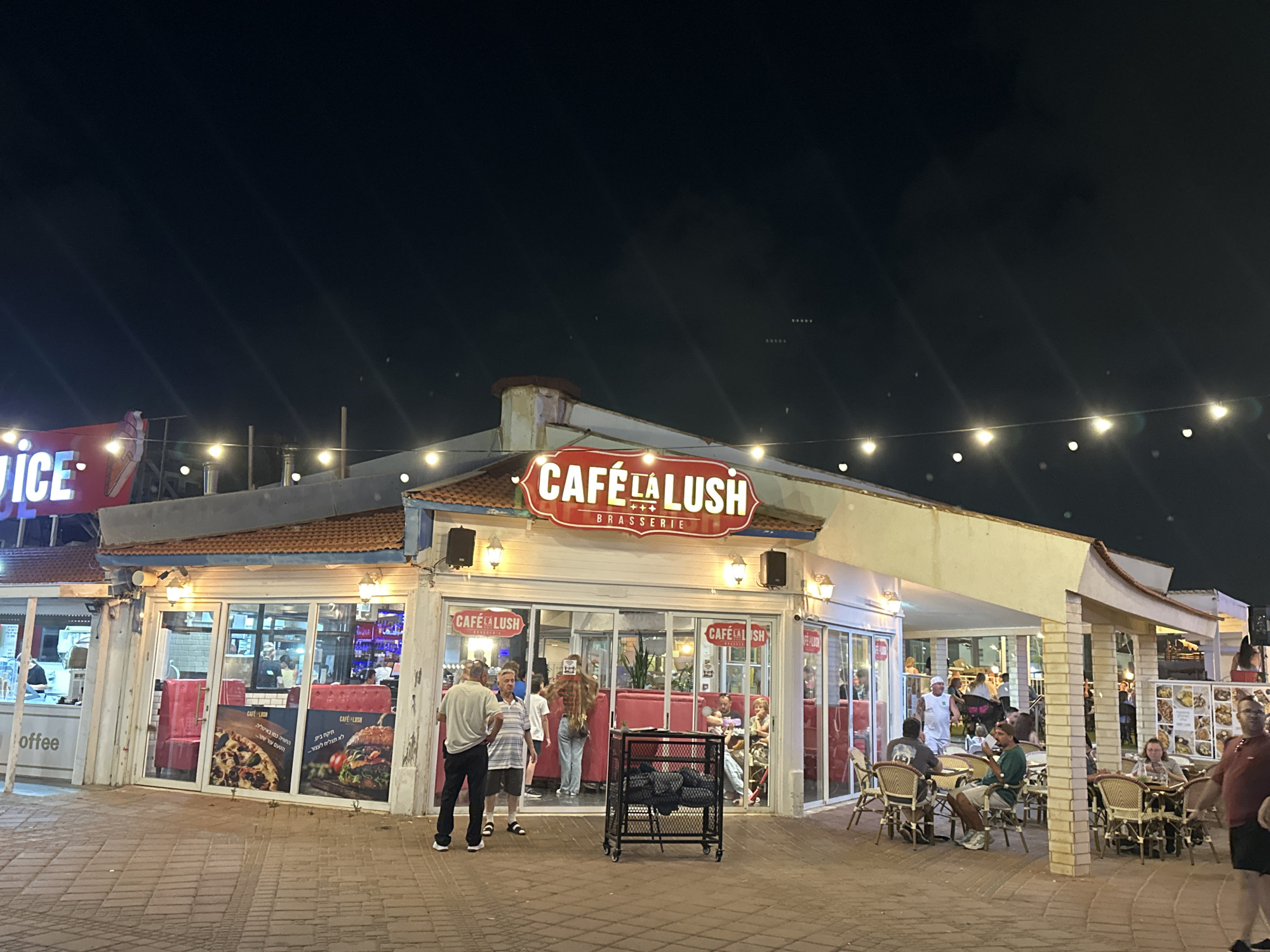 The image shows a street-level view of Cafe La Lush Brasserie, a restaurant and bar located in Nahariyya, Israel. It is nighttime, and the establishment is illuminated by string lights and interior lighting. The cafe features outdoor seating with tables and chairs, and a covered outdoor seating area. Several people are visible, some seated at tables and others standing. The facade of the cafe includes large glass windows displaying menu items, including pizza and a burger. The overall atmosphere appears to be lively and social.