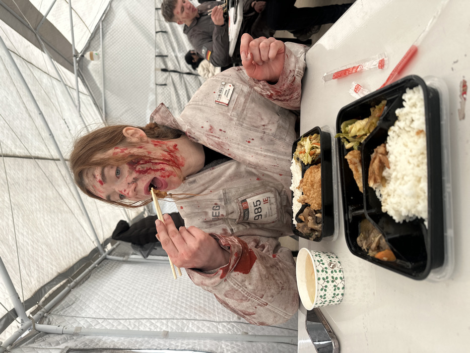 Inside a large white tent structure with insulated-appearing walls, a young adult female with reddish-brown hair is seated at a white table, consuming food. Her face and a light-colored jumpsuit are extensively covered in simulated blood. Patches on her jumpsuit include "VEG," "985 IE," and a name tag identifying "EMMA JORDAN." She uses chopsticks to guide food to her open mouth. On the table in front of her are two black, compartmentalized meal containers, each holding white rice, various meats (fried chicken, pork), and stir-fried vegetables. A paper cup containing soup and a silver smartphone are also on the table. In the background, a male individual wearing a dark jacket with a German flag patch is visible, also eating with chopsticks. This scene is situated in the Huangdao District, China.