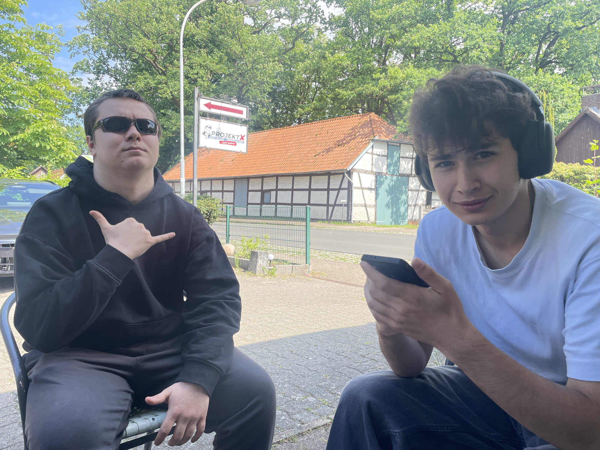 Two young men are sitting outside on a patio or sidewalk. The man on the left wears sunglasses and a black hooded sweatshirt. He's holding his hand up in a "hang loose" or "shaka" gesture. The man on the right is wearing headphones and holding a smartphone.
Behind them, across a street or road, is a half-timbered building with a red tile roof, similar to traditional German architecture. There is a sign near the building that says "PROJEKT X." The background suggests a rural or suburban setting with trees and foliage.
The location is Wienhausen, Germany, based on the signage in the background. The setting appears to be outdoors on a paved area near a building.