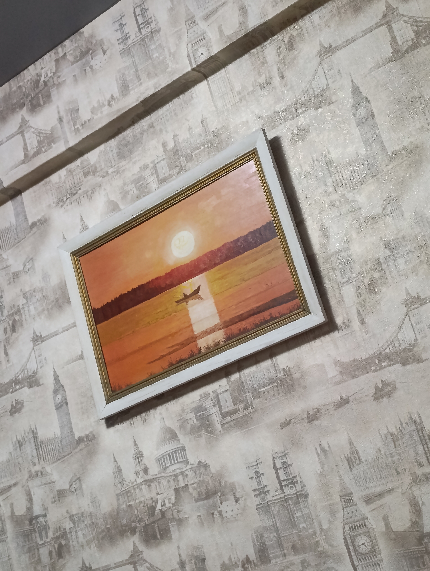 A framed painting hangs on a wall covered in London-themed wallpaper. The painting depicts a sunset over water with a small boat silhouetted in the foreground. The frame is white with a gold inner border. The wallpaper features grayscale images of London landmarks like the Tower Bridge, Big Ben, and St. Paul's Cathedral. The photo was likely taken indoors. There is no information on the location or specifics of the Krasnoyarsk, Russia location, and no other subjects, objects, actions or interactions are discernible in this image.