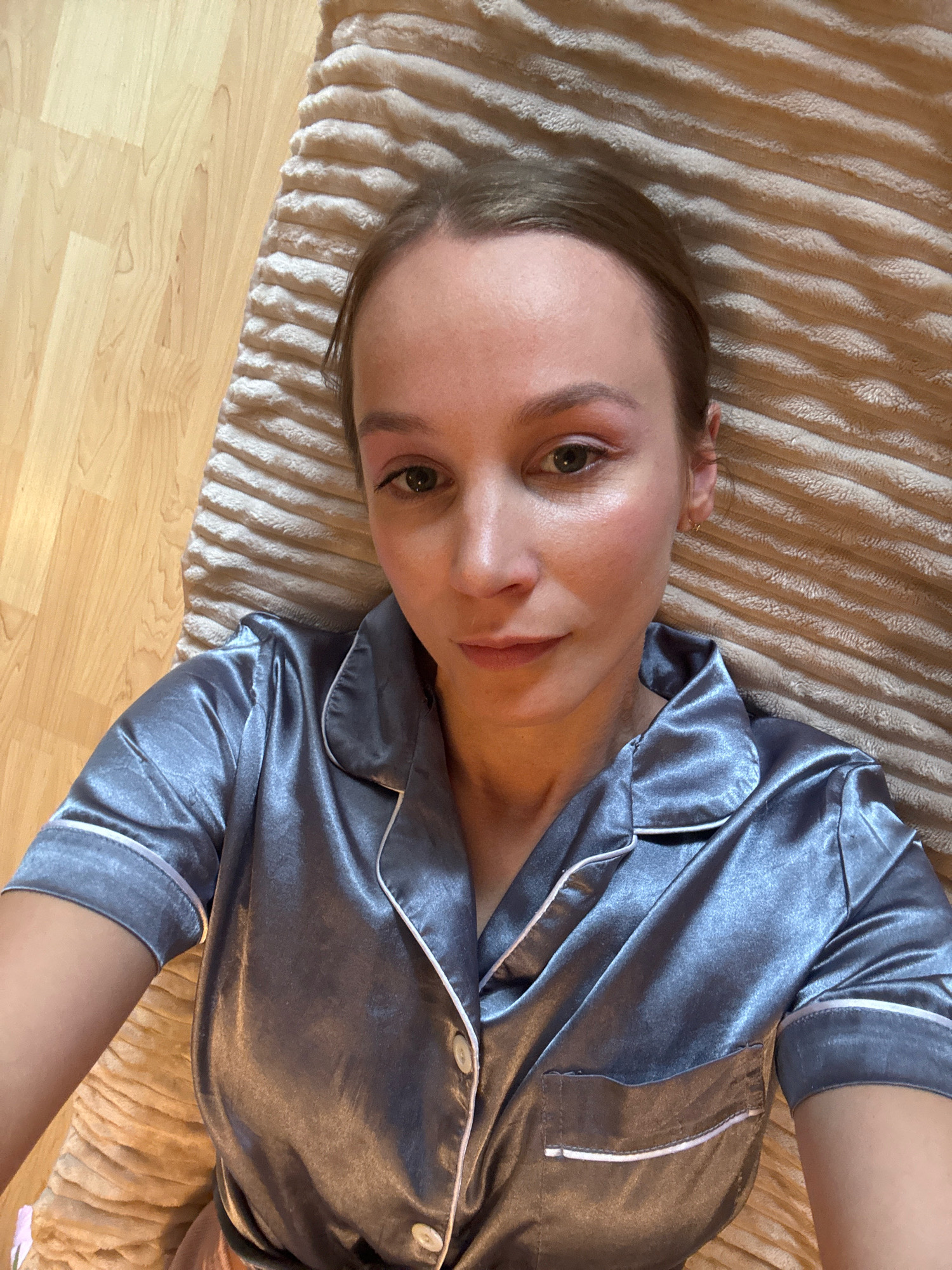 A young woman is lying on a rug on a wooden floor, looking at the camera. She is wearing a gray satin pajama top with white trim. Her hair is pulled back. The rug has a ridged texture and is beige in color. The wooden floor is light brown with visible grain. The image appears to be a selfie taken indoors. The location is not specified in the image or its context.