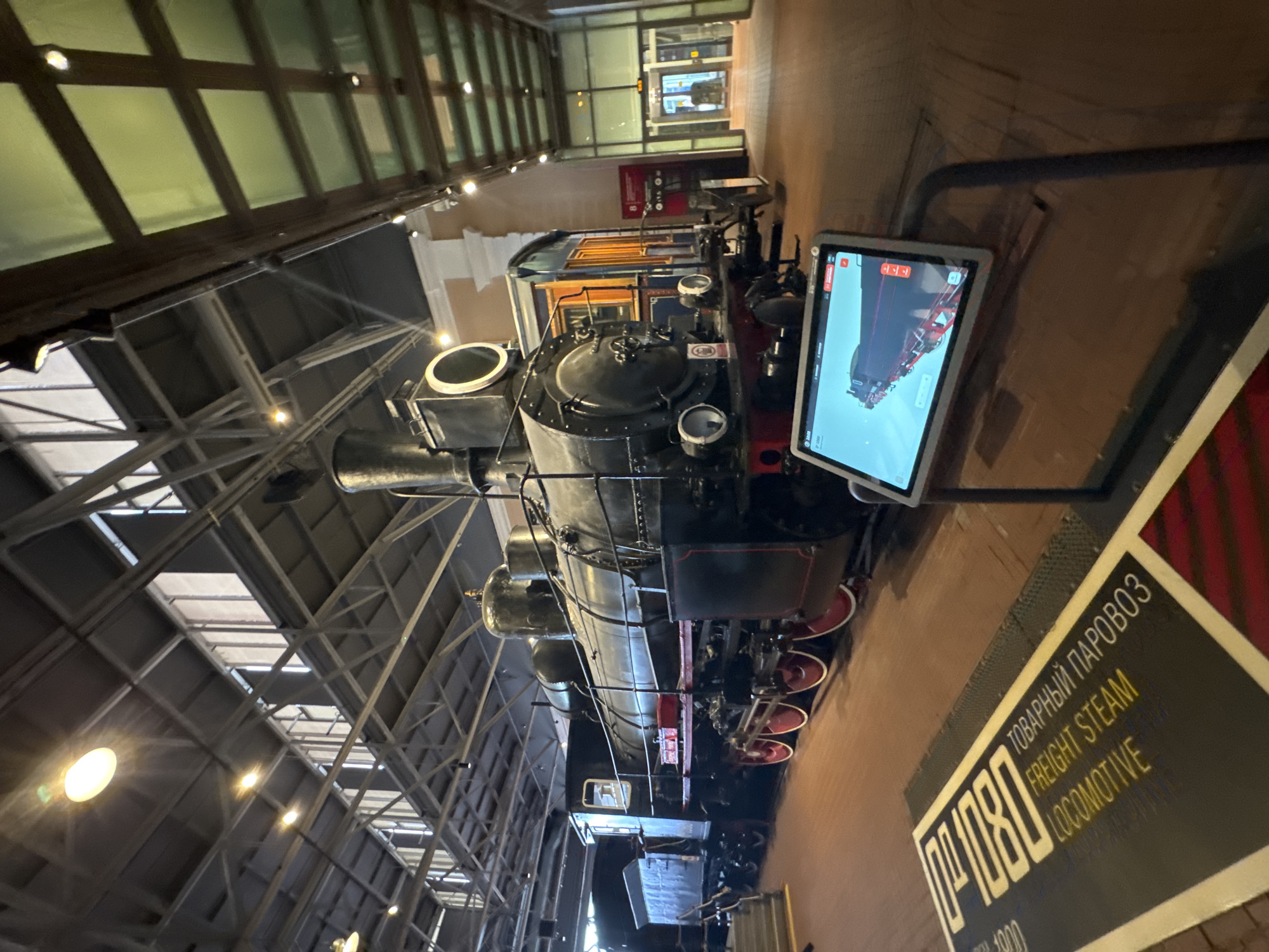 An antique black steam locomotive, identified by a nearby plaque as "№ 1080 Freight Steam Locomotive," is displayed indoors within a museum or exhibition hall. The locomotive features red-rimmed wheels, a large smokestack, and a tender car. To the right of the locomotive, a tablet displays a 3D rendering of the same train. The environment appears to be a spacious building with a high ceiling supported by metal beams and industrial lighting fixtures. The floor is made of wood planks. The text on the plaque is in both Russian and English. The scene is well-lit and suggests a historical exhibit about railways in Saint Petersburg, Russia.