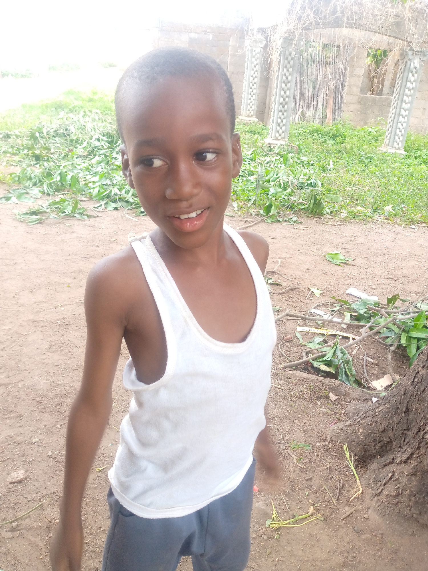 A young boy wearing a white tank top and grey shorts stands on brown soil. He is looking to his left with a slight smile. The background shows scattered green vegetation, tree branches, and a partially constructed building with decorative pillars. The image appears to be taken outdoors in a somewhat rural setting. It is not possible to verify the location in the city of Akwanga, Nigeria.