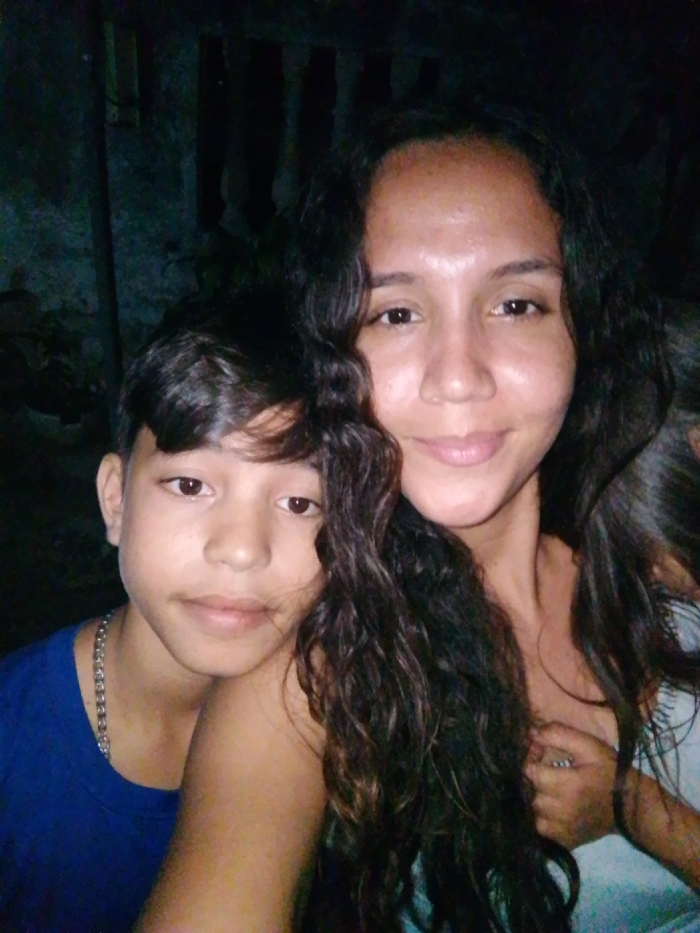 A woman with long, dark, curly hair smiles gently. Beside her, a younger person with short, dark hair and a chain necklace looks at the camera. The lighting is low, casting shadows in the background, which appears to be a dimly lit outdoor setting with hints of foliage. The woman is wearing a light-colored top, and the younger person is wearing a blue t-shirt. The overall tone is casual and familial.