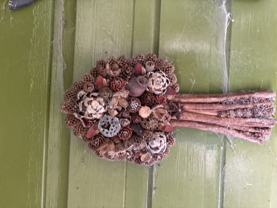 A decorative, tree-shaped natural arrangement is vertically mounted against an olive-green horizontal wooden paneled surface in the city of Stiperstones, United Kingdom. The item measures approximately 40 cm in height and consists of a dense, rounded top section composed of various dried natural elements, including numerous small pine cones, larger textured seed pods, and stylized flower-like components, some appearing silvery and others reddish-brown. The lower section forms a bundled "trunk" constructed from vertical cinnamon sticks or similar reddish-brown wood bundles, with several star anise pods embedded near the top of this section. The entire arrangement and the adjacent green paneling exhibit visible accumulations of fine dust and white cobwebs, indicative of prolonged exposure to ambient conditions without recent cleaning. No individuals, actions, or direct interactions are depicted.