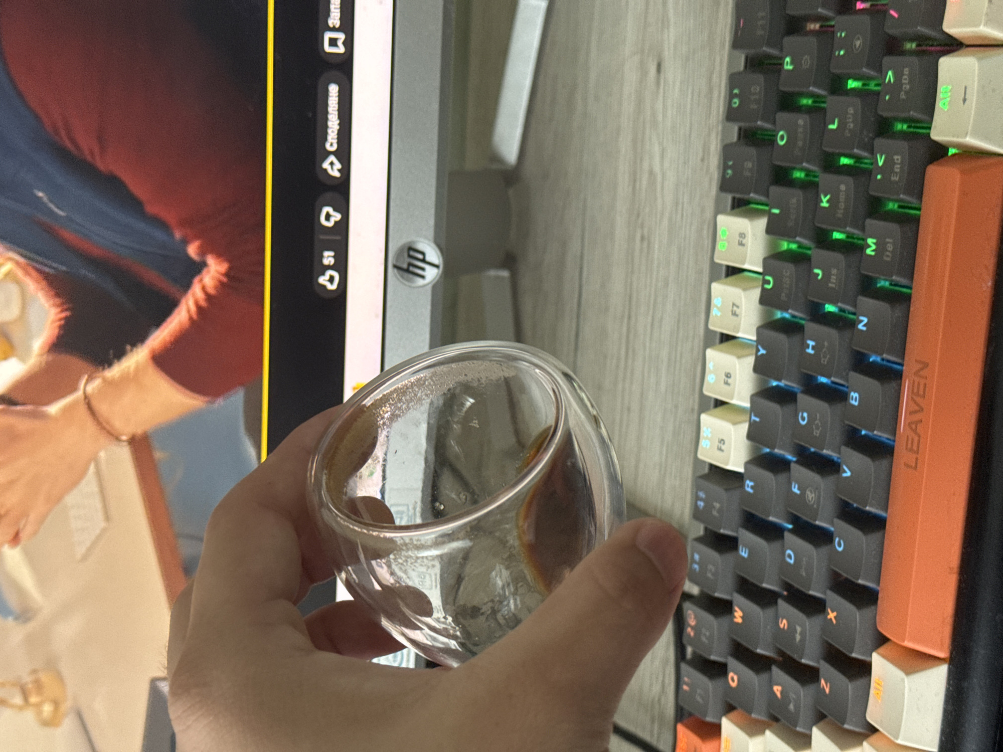Here's a detailed description of the image:
**People:** A hand holding a glass with a dark liquid (likely coffee or espresso) is visible. Another hand is partially visible on the computer screen.
**Subjects:** The primary subjects are the hand holding the glass, the glass itself, and the keyboard. The computer screen in the background is also a subject.
**Objects:** The key objects include a double-walled glass cup with a dark liquid, a computer monitor (possibly an HP brand as indicated by the logo), a mechanical keyboard (branded "LEAVEN"), and some computer screen elements like a like and share button.
**Actions:** The main action is holding the glass.
**Interactions:** The glass is being held near the keyboard and the computer monitor. The computer is playing a video, where someone cooks.
**Environment:** The scene appears to be indoors, likely on a desk or table. The keyboard and computer monitor indicate a workstation setup. The background has a grey wooden pattern that could be a desk or a floor.
**Location:** There is not enough information in this image to determine if this is Varna, Bulgaria.