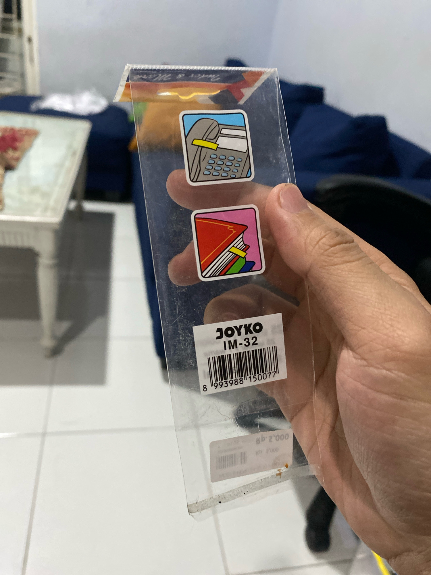 A person's hand holds a clear plastic packaging containing two stickers and a product label. The top sticker depicts a calculator. The bottom sticker shows a stack of colorful books. The product label below reads "JOYKO IM-32" and has a barcode. The background shows a blurry indoor environment with a white tiled floor, a white table with a patterned object on it, and a dark blue sofa. The image appears to be taken in a residential setting. The city of Klurak, Indonesia is not explicitly visible in the image, but the presence of Indonesian text on a small price tag below the main label (which reads "Rp. 2.000") suggests the location.