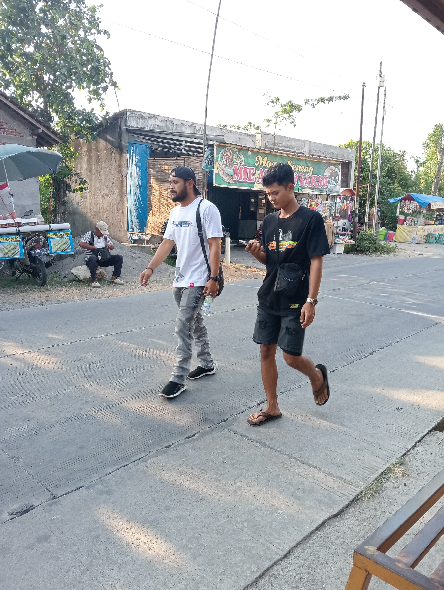 Two young men are walking on a paved street in front of buildings with a sign that reads "MIE AYAM BAKSO" in Tanjunganyar, Indonesia. The man on the left is wearing a white t-shirt, grey jeans, and black sneakers. He has a black baseball cap on and a black bag slung over his shoulder. He is holding a bottle of water and appears to be smoking. The man on the right is wearing a black t-shirt, black shorts, and flip-flops. He has a small black bag across his chest and is looking at his phone. In the background, a man in a striped shirt and cap is sitting on a rock next to a motorcycle with a food cart. Another food cart with a blue canopy is visible further down the street. Trees and power lines are also present in the scene. The lighting suggests it is daytime.