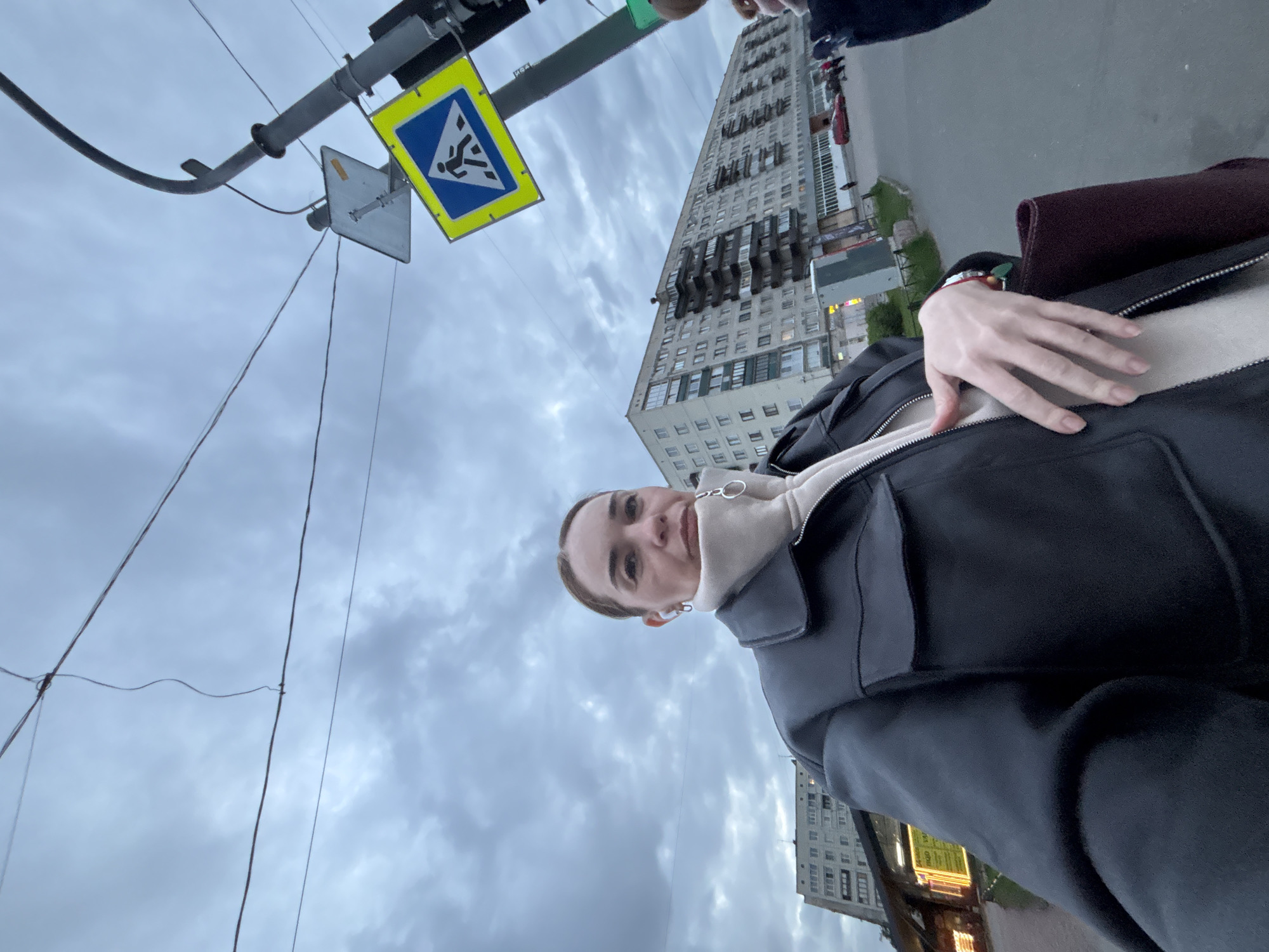 A low-angle, canted selfie captures a fair-skinned female with light brown hair pulled back, wearing earbuds, a high-necked tan top, and a black jacket. She is looking at the camera with a slight smile, her left hand resting on her jacket.
Behind her, a pedestrian crossing sign is visible, indicating the location is near a crosswalk. In the background, a tall, Soviet-era apartment building stands under an overcast sky with heavy clouds. A segment of a city street is visible on the right, along with the upper stories of a commercial building with illuminated signage.
The location is the city of Unknown, Unknown.