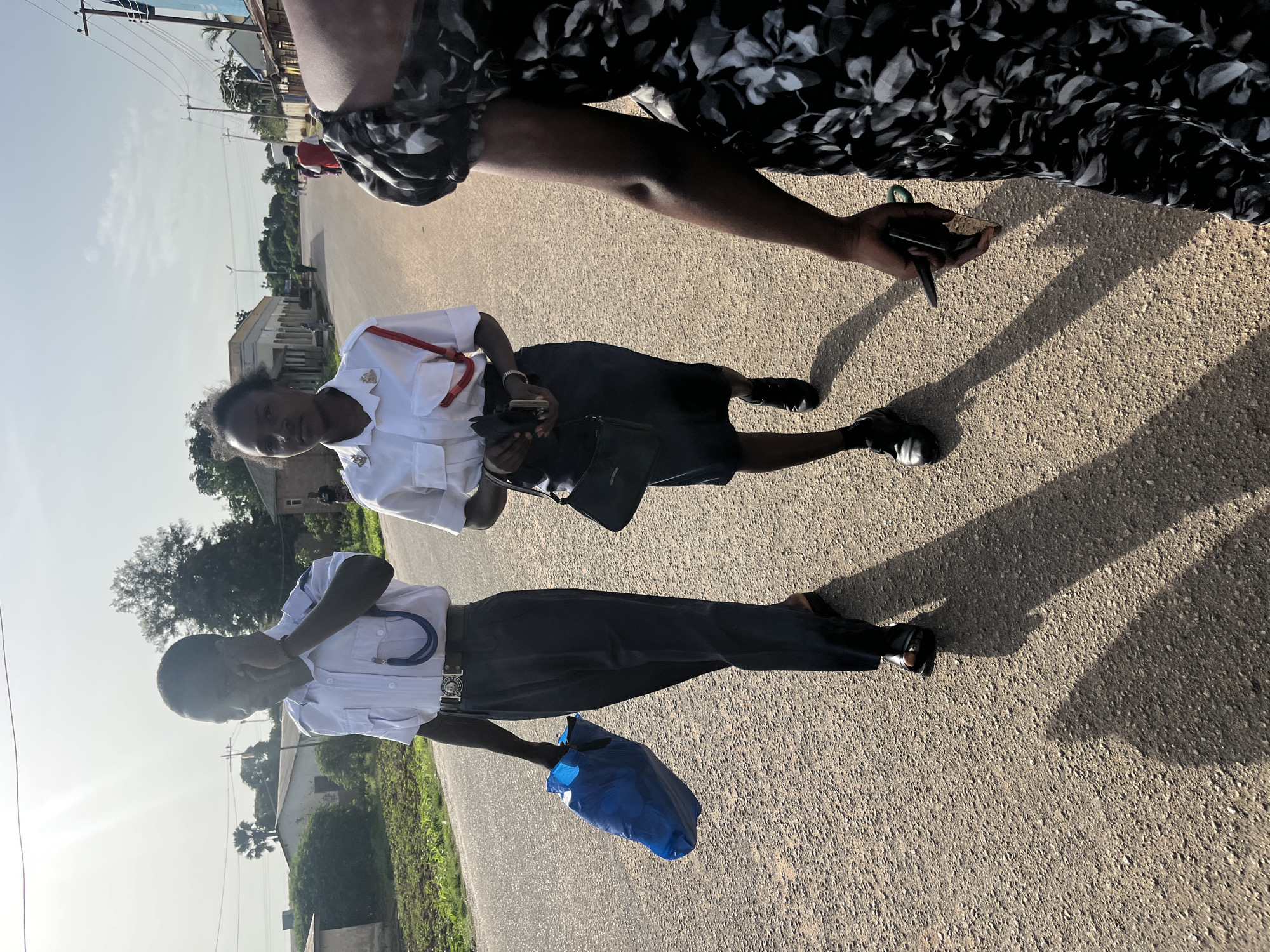 In Bukuru, Nigeria, two individuals in uniform walk down a paved road under a bright sky. The person on the left, appearing to be a male, wears a white short-sleeved shirt with epaulets and a dark blue tie or sash with a blue string hanging down. He has dark skin and short hair, and is holding a bright blue plastic bag in his left hand while appearing to speak on a mobile phone in his right. He wears dark blue trousers and black sandals. The person on the right, appearing to be a female, wears a similar white short-sleeved shirt with red accents on the epaulets and a dark blue skirt. She has dark skin and dark, pulled-back hair. She carries a small black purse on her left arm and holds a black mobile phone in her right hand. She wears black shoes. A third person, partially visible on the right, wears a black and white patterned dress and holds a dark object, possibly a phone, in their right hand. The road is bordered by vegetation and buildings on either side, with power lines visible overhead. The sun casts long shadows on the ground, indicating it is likely late afternoon.