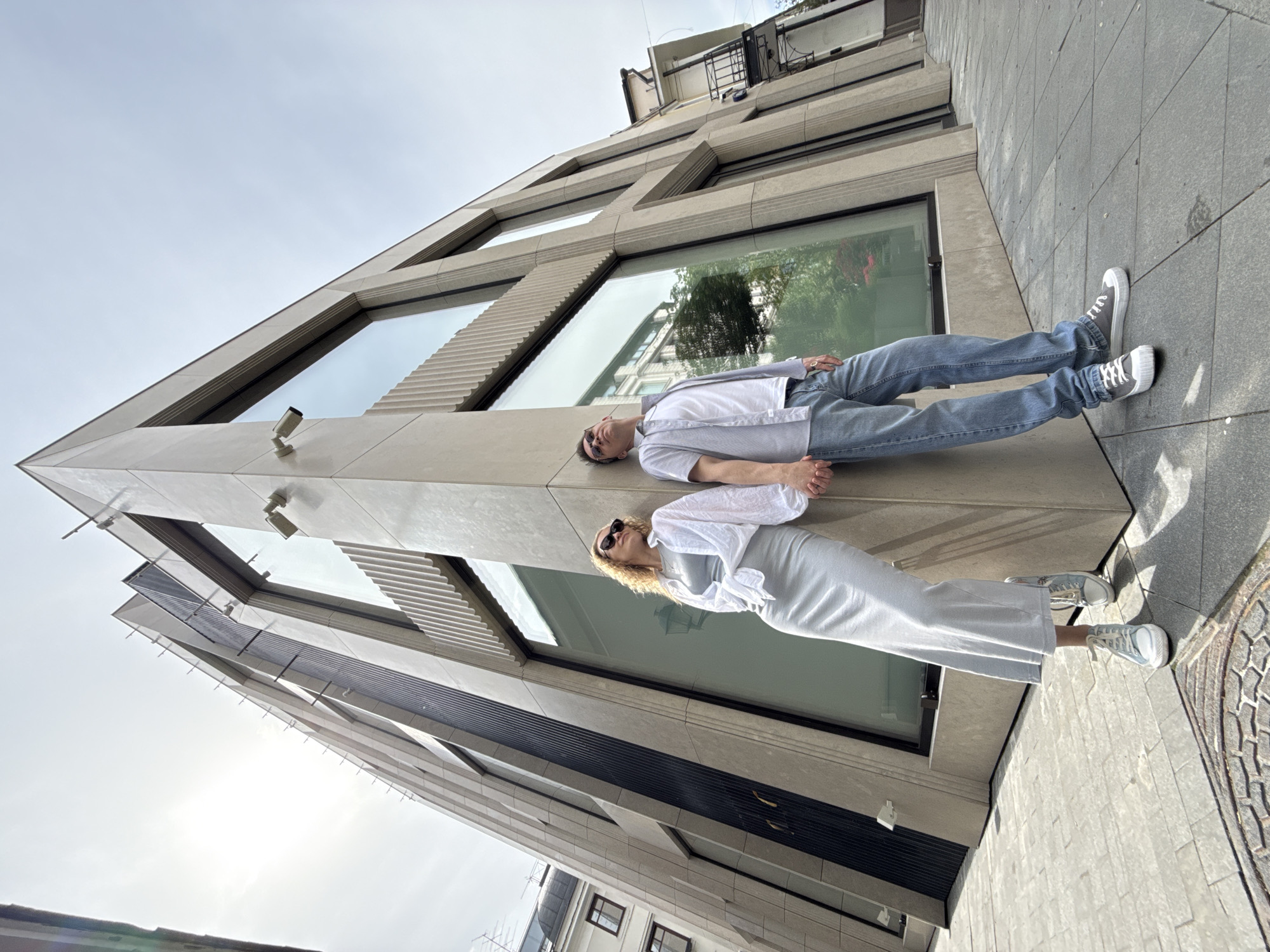 A low-angle, tilted perspective captures a man and a woman seemingly lying on the side of a building. The man is dressed in a white t-shirt, a light blue button-up shirt, and blue jeans, with grey sneakers. He wears sunglasses. The woman wears a light-colored dress or skirt and blouse, with a white long-sleeve top and light-colored sneakers. She wears sunglasses as well. They appear to be posing against the building's exterior, which has large windows and geometric design elements, including security cameras. The background includes the sky and parts of other buildings, suggesting an urban environment. The location is Moscow, Russia.