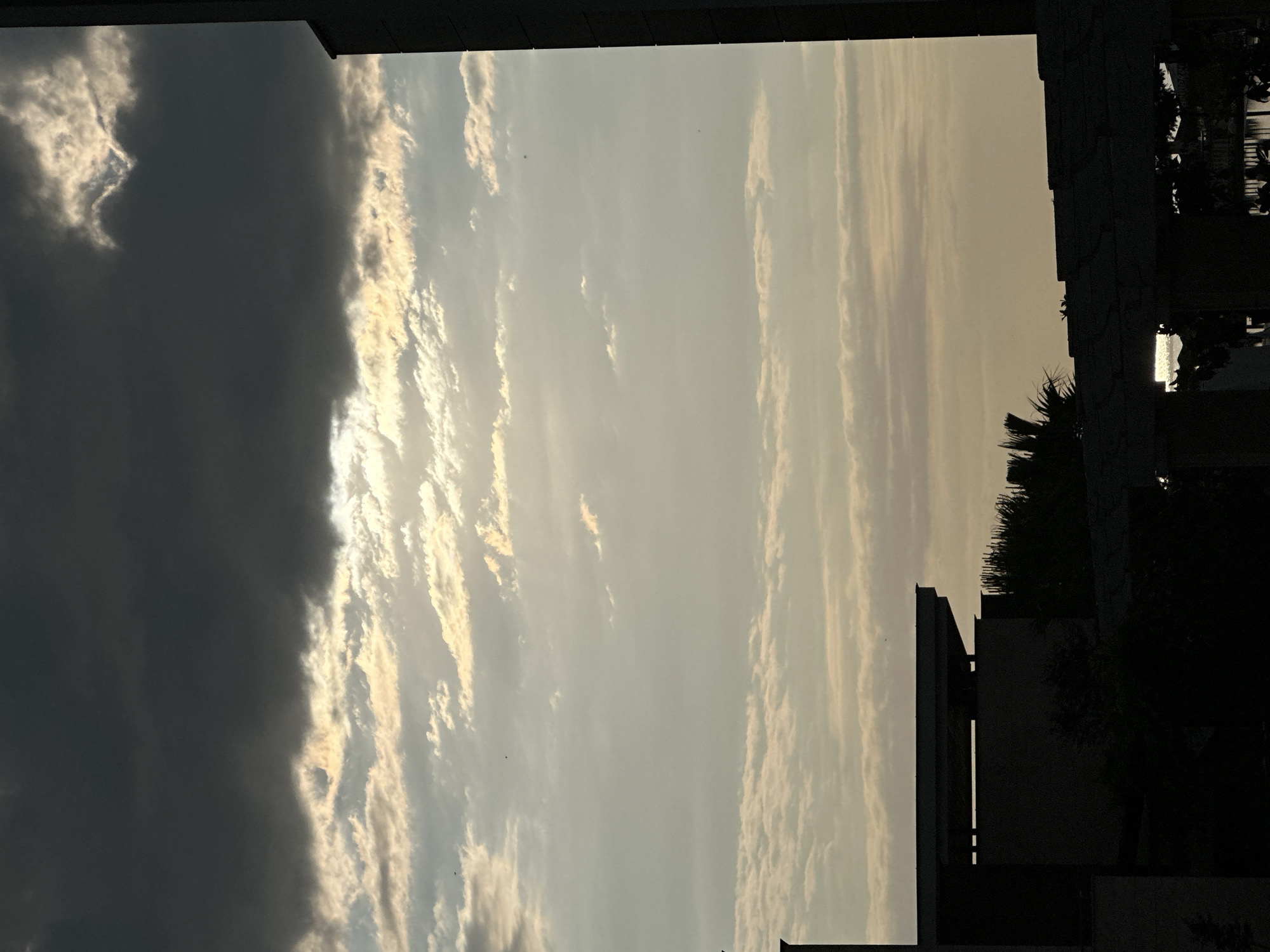 The image shows a view of the sky with clouds and the silhouettes of buildings. The sky is a mix of dark grey and lighter, yellowish-white clouds. The sun is partially visible through the clouds, creating a bright halo effect. In the foreground, the dark silhouettes of several buildings are visible, including one tall, dark rectangular structure on the right side of the image. The buildings appear to be modern architecture. The overall scene suggests it might be late afternoon or early evening, given the lighting conditions and cloud formations. The image does not contain any people or specific details that would allow for identification of the location within the city of Sozopol, Bulgaria.