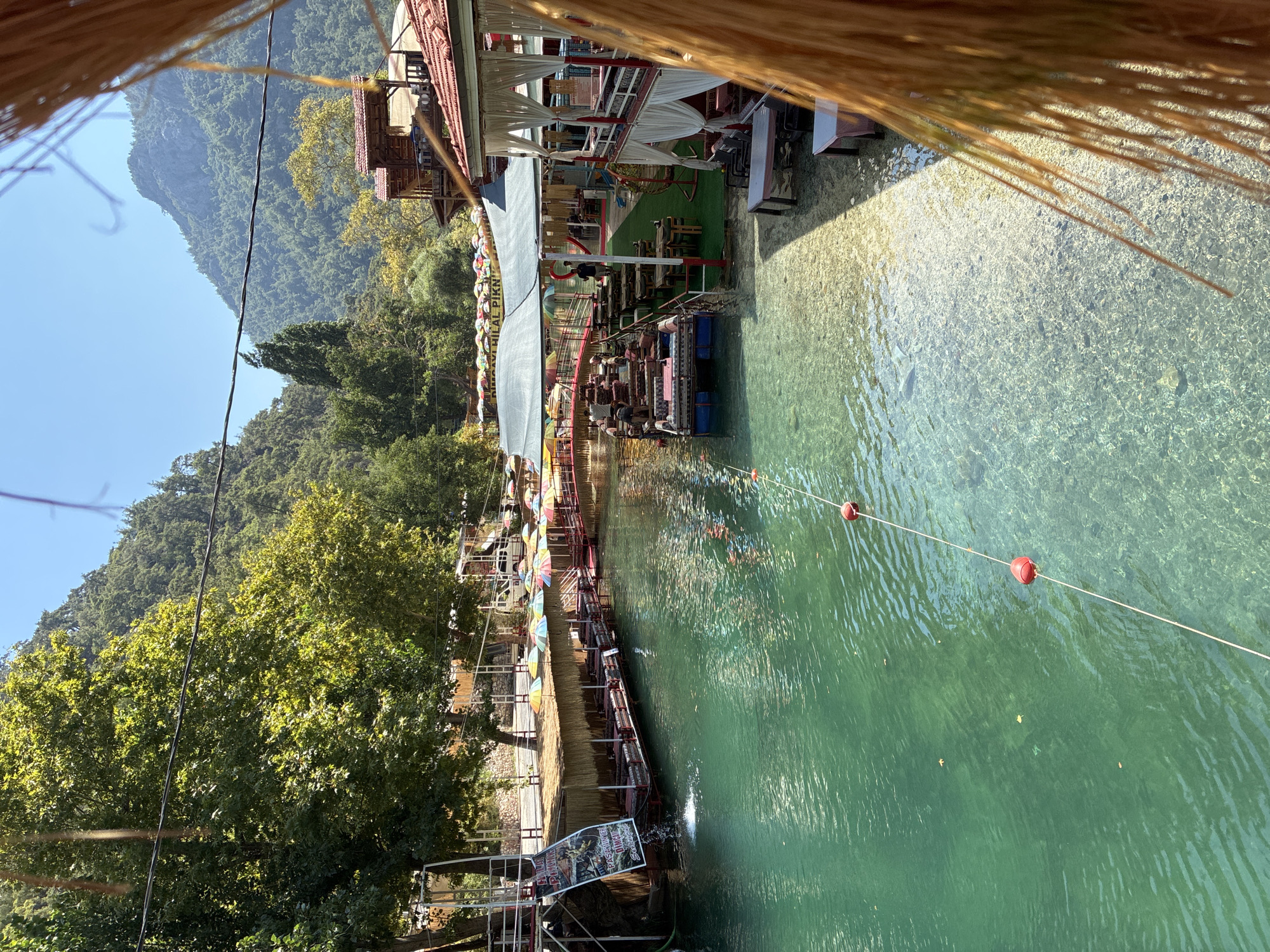 This image depicts a scenic riverside restaurant in Alanya, Turkey, characterized by its clear, turquoise water and lush green surroundings. The restaurant features several seating areas, including platforms suspended over the water and covered gazebos, all furnished with cushions and tables. Colorful umbrellas provide shade over some of the dining spaces. The water is shallow and transparent, allowing the rocky bottom to be visible. Several red buoys are tethered by a rope, marking a section of the water. In the background, steep, tree-covered mountains rise against a clear blue sky, indicating a natural, possibly rural, setting within or near Alanya. A few people are visible, some attending to the restaurant operations and others seated at the tables. The overall atmosphere suggests a tranquil and inviting dining experience amidst nature.