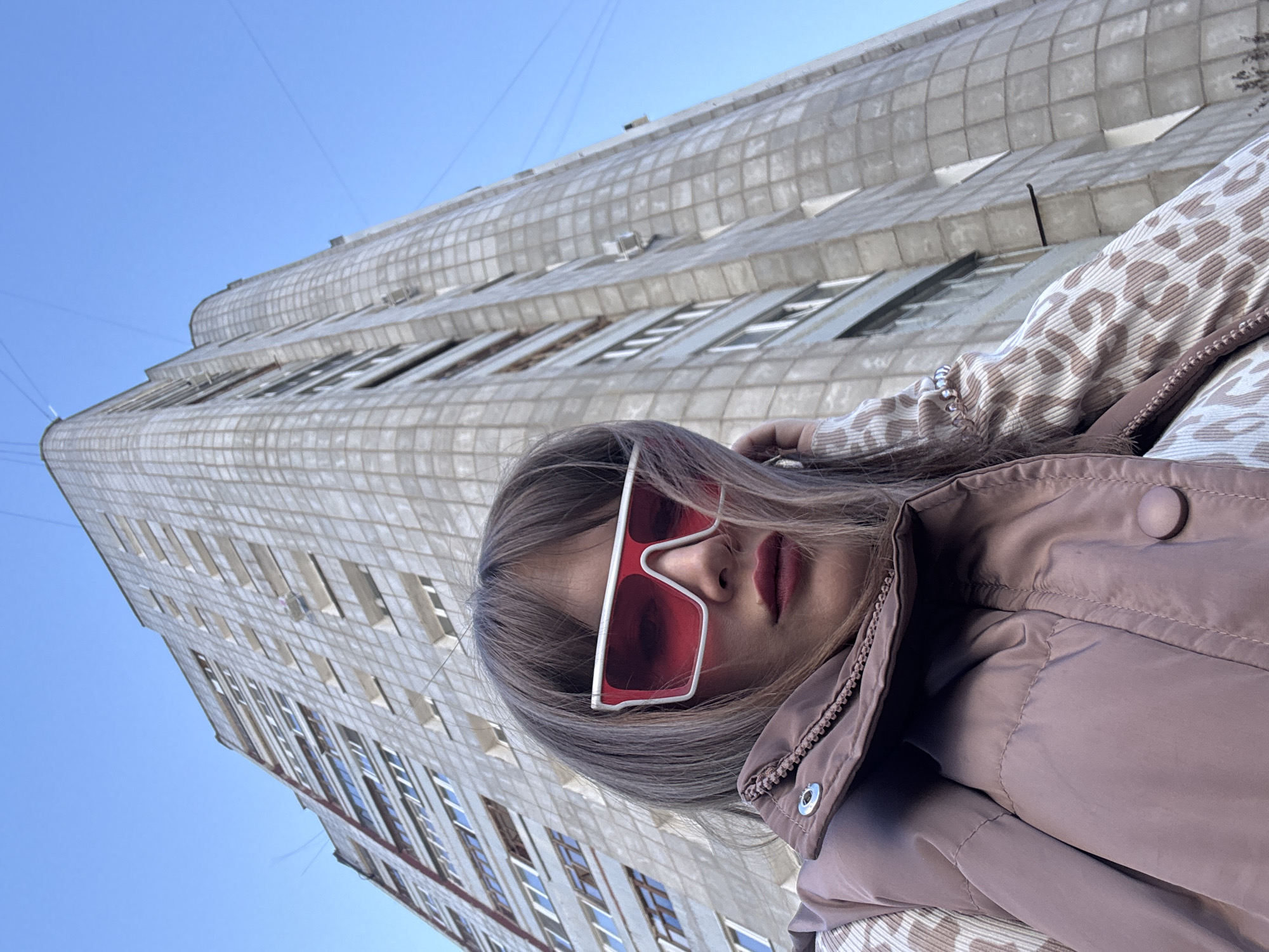 A low-angle, tilted outdoor shot depicts a woman wearing large, red-lensed white-framed sunglasses, a mauve jacket, and a beige and white patterned top. She appears to be taking a selfie. Her face is the primary focus, with a portion of a high-rise building as the backdrop. The building is made of light gray, grid-like blocks with numerous windows reflecting the blue sky. The sky is visible in the upper left corner and is a clear, light blue. The location is the city of Chaikovsky, Russia.