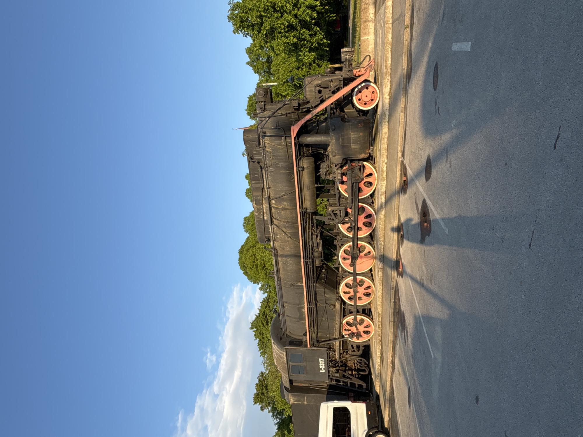 A large, black steam locomotive is prominently displayed outdoors, likely as a static exhibit. The train features several substantial driving wheels, notable for their red-orange spokes and white rims. White text reading "L-2317" is visible on the side of the locomotive's cab.
The scene is set alongside an asphalt road with a concrete curb. In the background, a line of dense green trees stretches beneath a clear, bright blue sky with a few scattered white clouds. Long shadows cast across the road suggest that the photograph was taken during either the early morning or late afternoon hours on a sunny day. Part of a white vehicle, possibly a van, is visible in the bottom foreground, to the right of the locomotive's front end.
The image depicts a historical piece of machinery, appearing to be on display in a public area or open-air museum in Tallinn, Estonia. There are no people visible, and no active events are taking place; the scene focuses solely on the stationary locomotive.