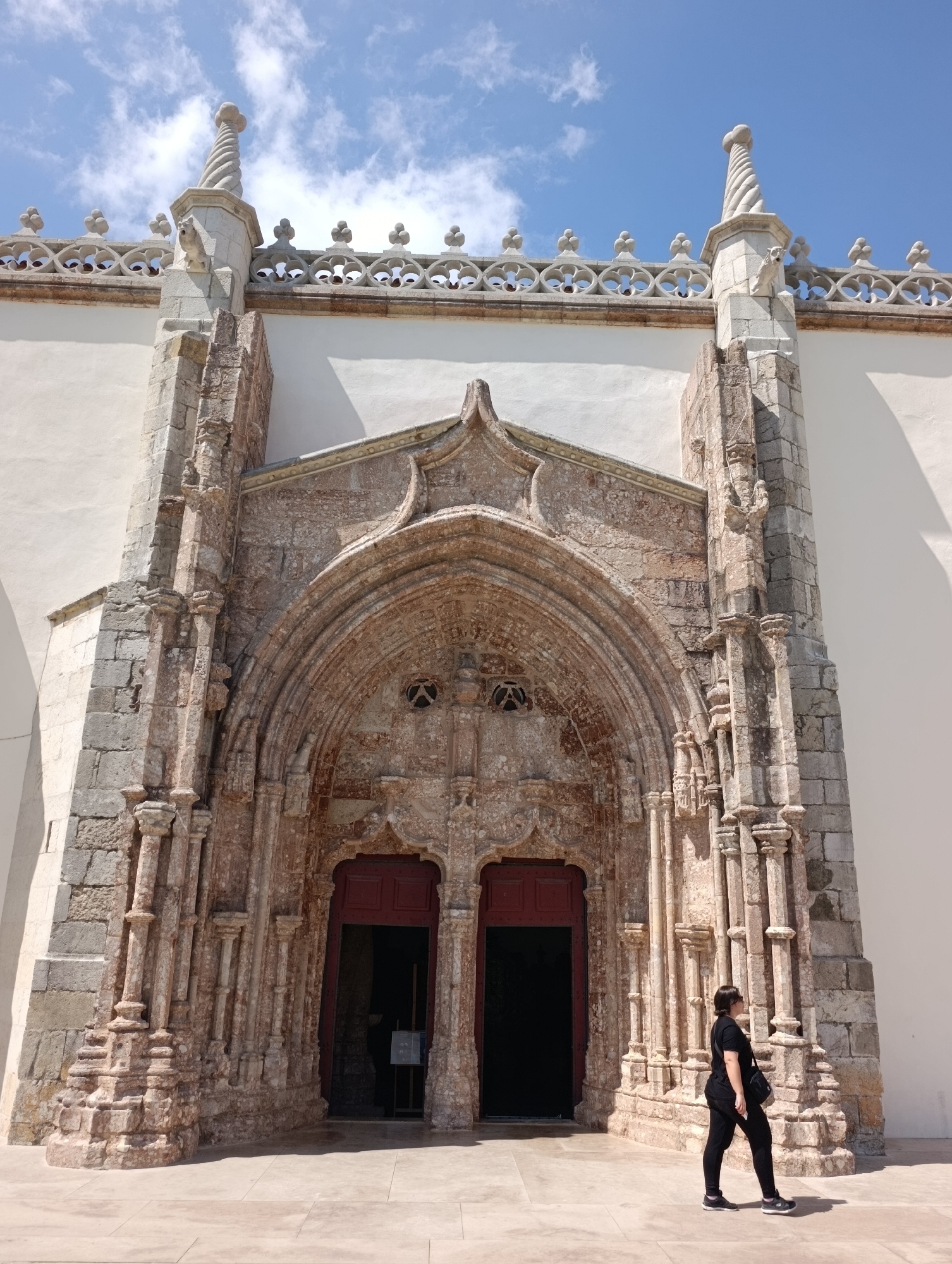 The image captures the exterior of the Church of Jesus of Setúbal (Igreja de Jesus de Setúbal), a National Monument located in Setúbal, Portugal. The church's ornate Gothic-Manueline portal dominates the foreground, featuring intricate stone carvings and arched doorways. A woman, dressed in black, stands to the right of the entrance, looking towards the building. The sky above is blue with scattered white clouds. The foreground consists of a paved courtyard. The overall scene is one of architectural grandeur set against a clear, bright day.