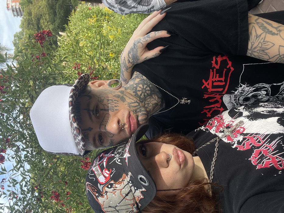 A young male and female are depicted in a close-up, chest-up selfie format, positioned outdoors in an urban park setting within Buenos Aires, Argentina. The background consists of lush green foliage, prominent red flowering trees (likely Ceibo trees, *Erythrina crista-galli*), and scattered yellow flowers. A light-colored, multi-story building is visible in the distant upper-right, under a partially cloudy sky.
The male, on the right, possesses extensive facial and neck tattoos, including a simulated band-aid below his left eye and the numbers "1312" on his right cheek. He has large ear gauges, a septum piercing, and two nostril piercings. He wears a white trucker hat with a leopard print brim and a black t-shirt featuring a stylized red "SLAVES" text graphic above a white and black illustration of a skull wearing a crown of thorns. A silver ball-chain necklace with a small silver cross pendant is visible. His right arm is around the female's shoulders, and his left arm is not fully visible, likely holding the camera.
The female, on the left, has reddish-brown hair partially visible beneath a black baseball cap adorned with white eyelets, and intricate silver and red-orange graphics that appear to depict an eagle or phoenix with text. She has a septum piercing and two nostril piercings. She wears a black t-shirt with a white and red graphic print. A chunky silver chain necklace with a silver cross pendant is visible around her neck. Her left hand, with long, black-painted fingernails, rests on the male's right shoulder. Both individuals are looking directly at the camera, with the female exhibiting slightly pouting lips.