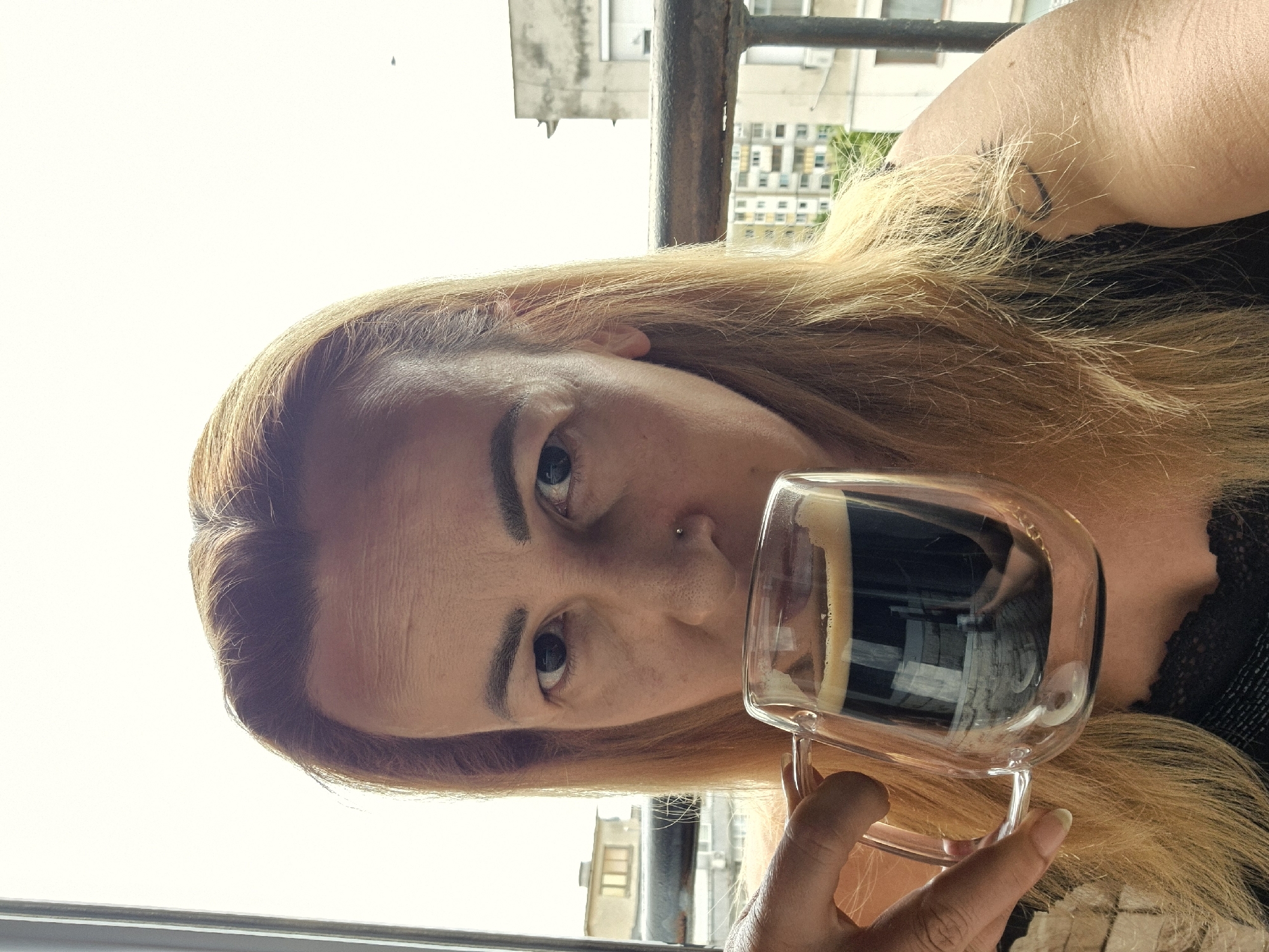 A medium-shot, eye-level, outdoor selfie shows a person with long blonde hair holding a clear glass mug filled with dark liquid and foam, likely coffee, up to their mouth. The person has dark eyebrows, a nose ring, and is wearing a black top with lace detailing. In the background, a metal railing is visible, and beyond that, apartment buildings and an overcast sky. The scene appears to be a balcony or similar elevated outdoor space in an urban setting.