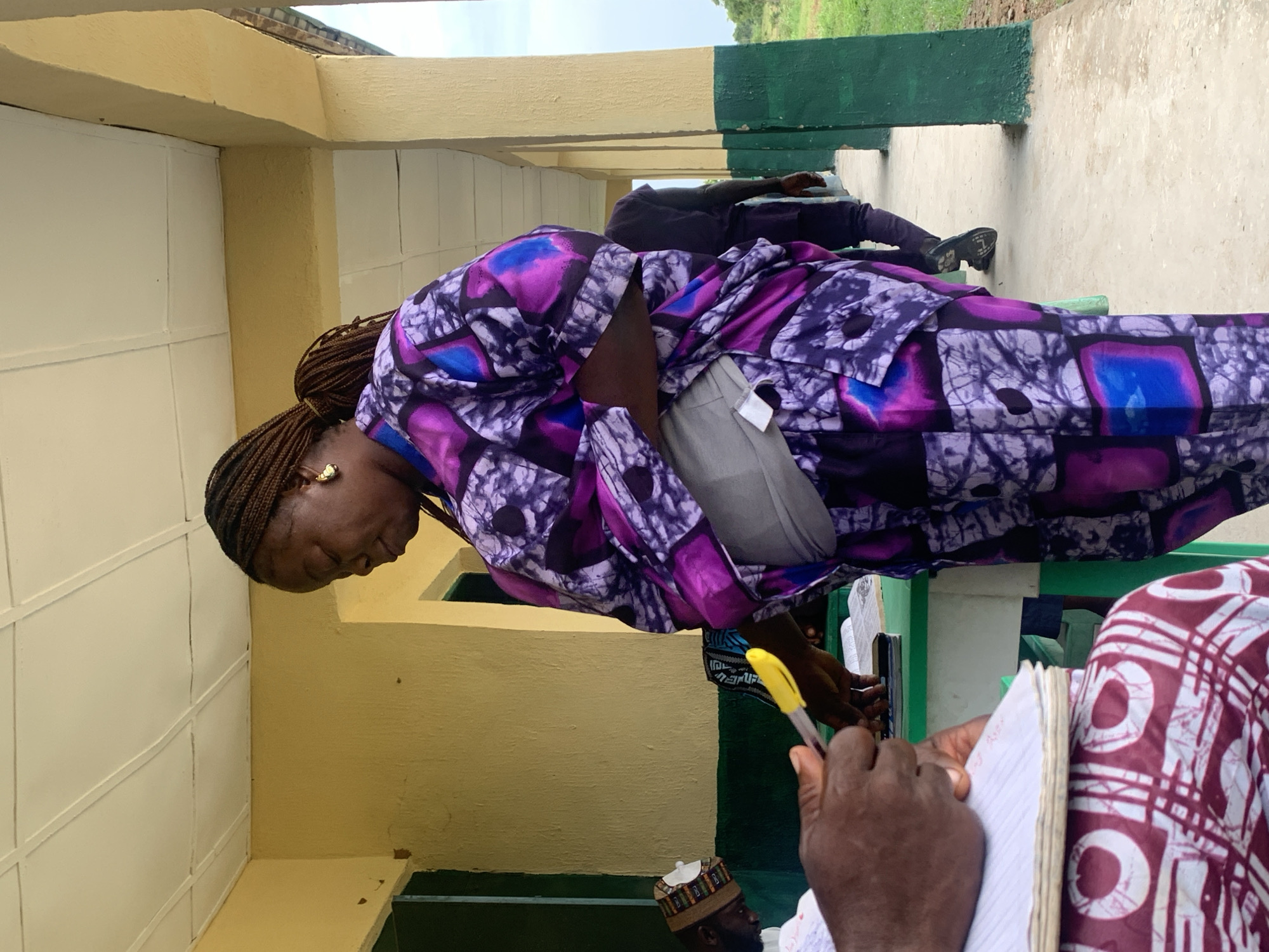 A woman in a purple patterned garment leans forward, appearing to examine or interact with something out of frame, while another person holds a notebook and pen in the foreground. The scene is set in what appears to be a corridor or covered walkway, with yellow and white painted walls. Another individual sits in the background, legs visible. The location is Tabu, Nigeria, based on the context provided.