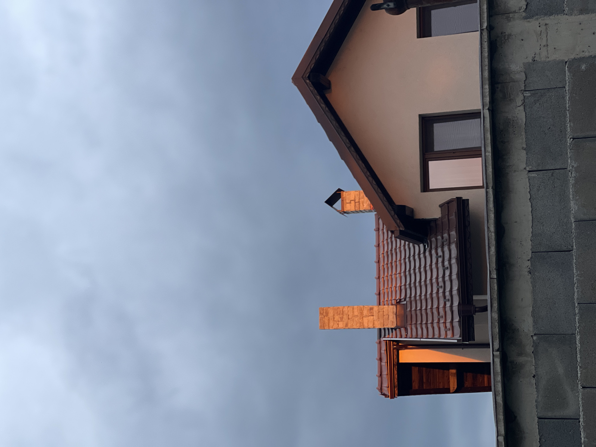 The image depicts a partial view of a house with a tiled roof, light-colored walls, and brown trim. The house has two visible windows. A chimney is present. The house appears to be under construction, as a gray brick wall stands adjacent to the house. The sky is overcast. It is not possible to determine the location in Sofia, Bulgaria from the image.