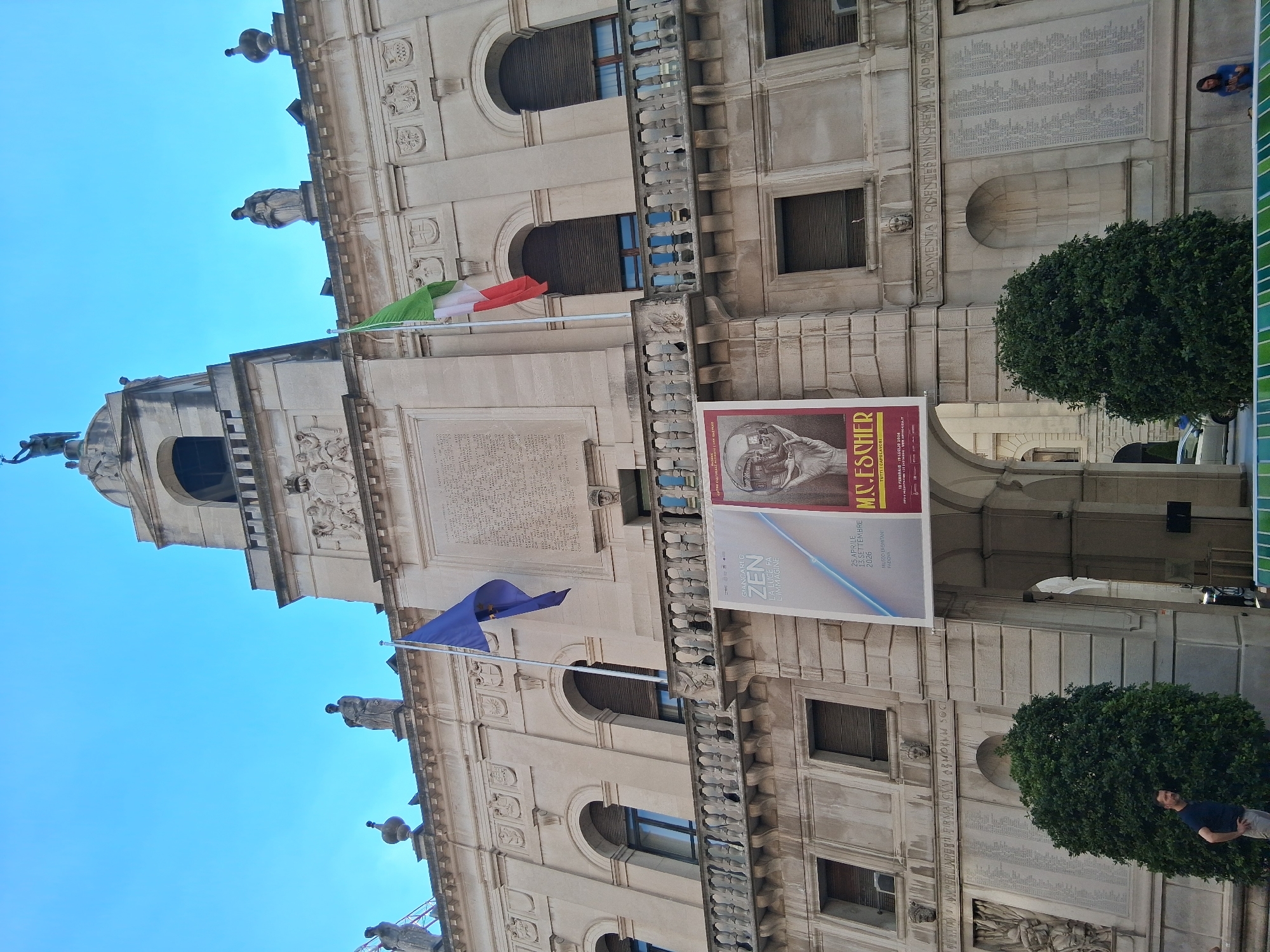 The image depicts the Palazzo Moroni, a historic building in Padua, Italy, under a clear blue sky. The facade of the building is made of light-colored stone and features classical architectural elements, including arched windows, balconies, and decorative sculptures. Two flags are visible: a blue flag with stars, likely the European Union flag, and the Italian flag. Large banners advertising an exhibition are hung on the building's facade. In the foreground, there are two well-maintained evergreen bushes. A man in a dark t-shirt and light-colored pants stands near the left bush, looking towards the building. Further back, a woman in a blue shirt is partially visible near the right side of the frame. The overall scene suggests a public space in front of a significant civic building.