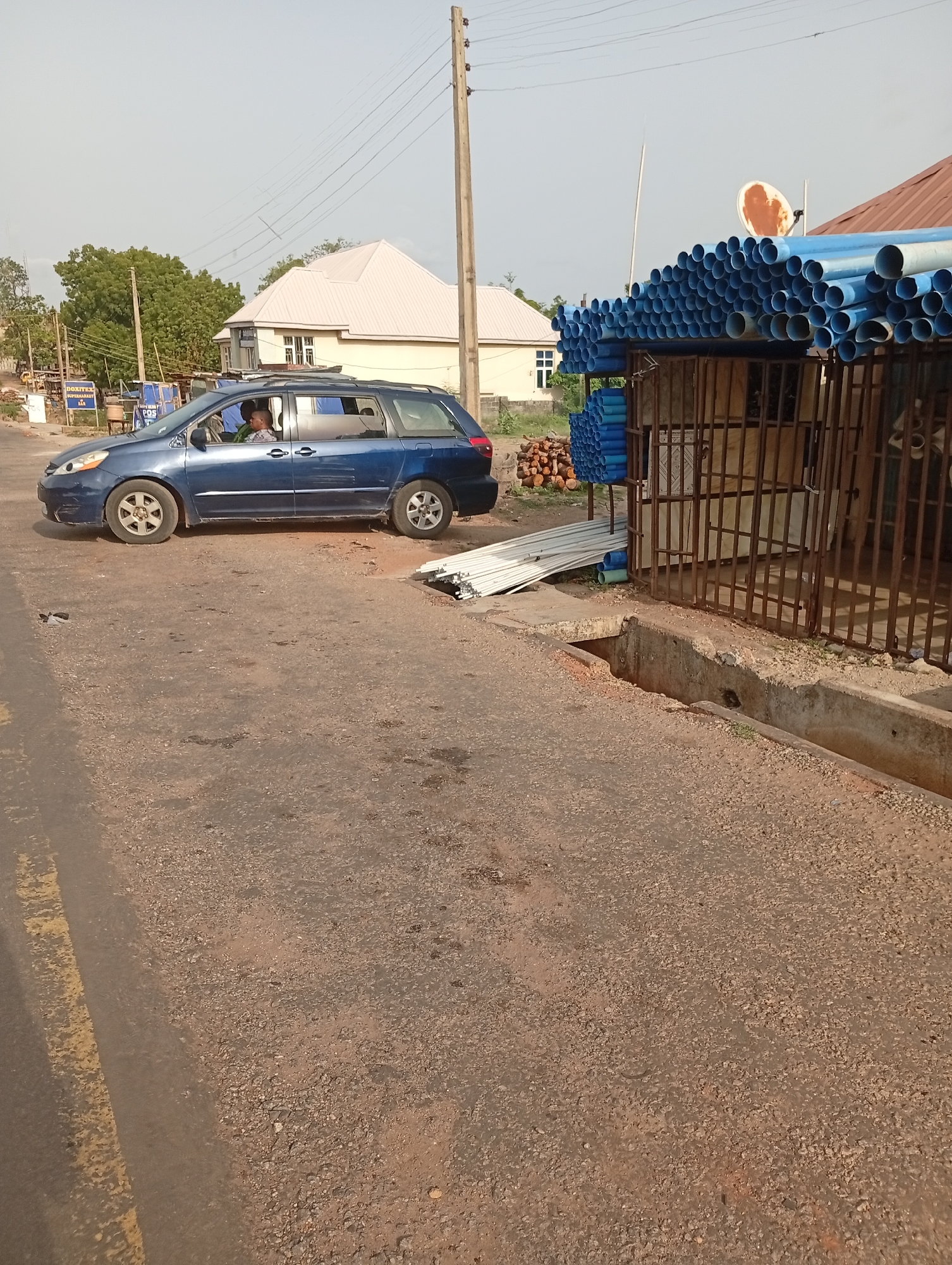 The image depicts a street scene in Adankolo, Nigeria. A blue minivan is parked on the side of the road. The road surface is paved, but in poor condition. Next to the road is a drainage ditch covered with a concrete slab and a grate. A small structure with a metal frame is laden with stacks of blue PVC pipes. A satellite dish sits atop this structure. In the background, a beige building with a white roof can be seen, along with a utility pole.