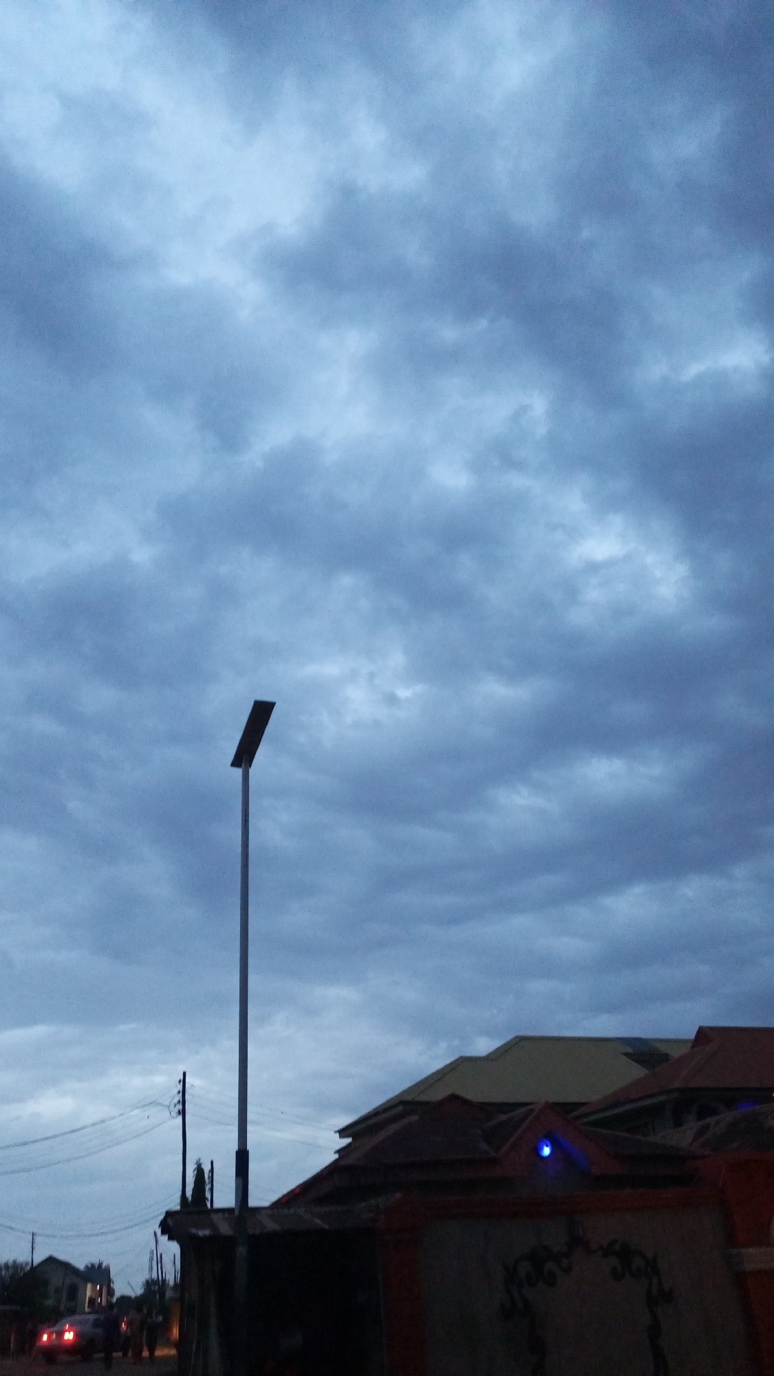 The image captures an overcast outdoor scene, likely during dusk or dawn, in Sabon Tasha, Nigeria. The sky is dominated by dense, dark grey clouds, creating a dim and muted atmosphere. A tall, slender lamppost, appearing to be a solar street light with a dark, rectangular panel at its top, stands prominently in the center-left of the frame, reaching towards the cloudy sky.
Below the lamppost and to the left, utility poles and power lines are visible against the gloomy light. In the foreground and midground, various building rooftops are seen, featuring shades of dark red, brown, and lighter beige. A distinctive detail is a small, glowing blue light or decorative element on the peak of one of the darker roofs.
At the bottom left, a street or road is discernible, where the faint outlines of a vehicle (possibly with illuminated taillights) and several indistinct human figures can be seen, suggesting general street activity. The overall impression is one of a quiet, transitional time of day in a residential or semi-urban environment, characterized by the heavy cloud cover and the emerging artificial light of the lamppost and the blue architectural detail. No visible text is present in the image.