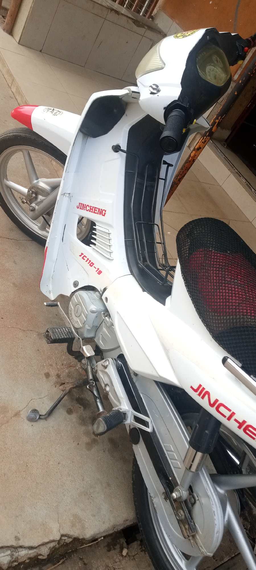 A white Jincheng JC110-19 motorcycle is parked on a concrete surface. The motorcycle has red Jincheng lettering, black handlebars, and a black and red mesh seat cover. The front of the motorcycle is slightly damaged. The location is suggested to be Jos, Nigeria.