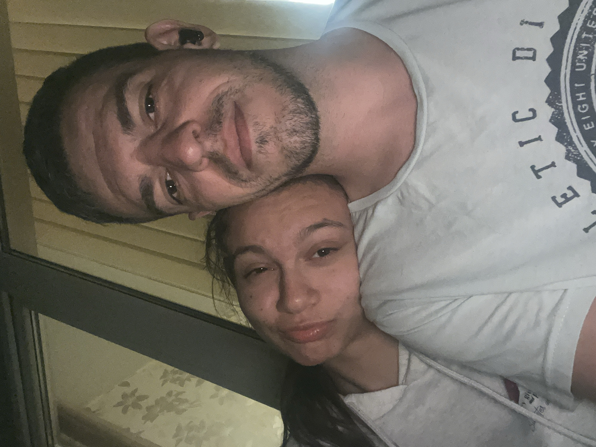 In this close-up selfie, a couple is resting indoors, with the woman's head on the man's shoulder. They are positioned in front of a window with floral patterned curtains, suggesting a residential setting. The man, in the foreground, has short dark hair and stubble, wearing a light-colored t-shirt with text on it. The woman, leaning on him, has long dark hair and appears to be wearing a light-colored hooded garment. Their relaxed posture indicates intimacy and comfort. The lighting suggests it might be evening or a dimly lit room. The presence of an earbud in the man's ear is also noted. No specific landmarks in Sofia, Bulgaria, are identifiable.