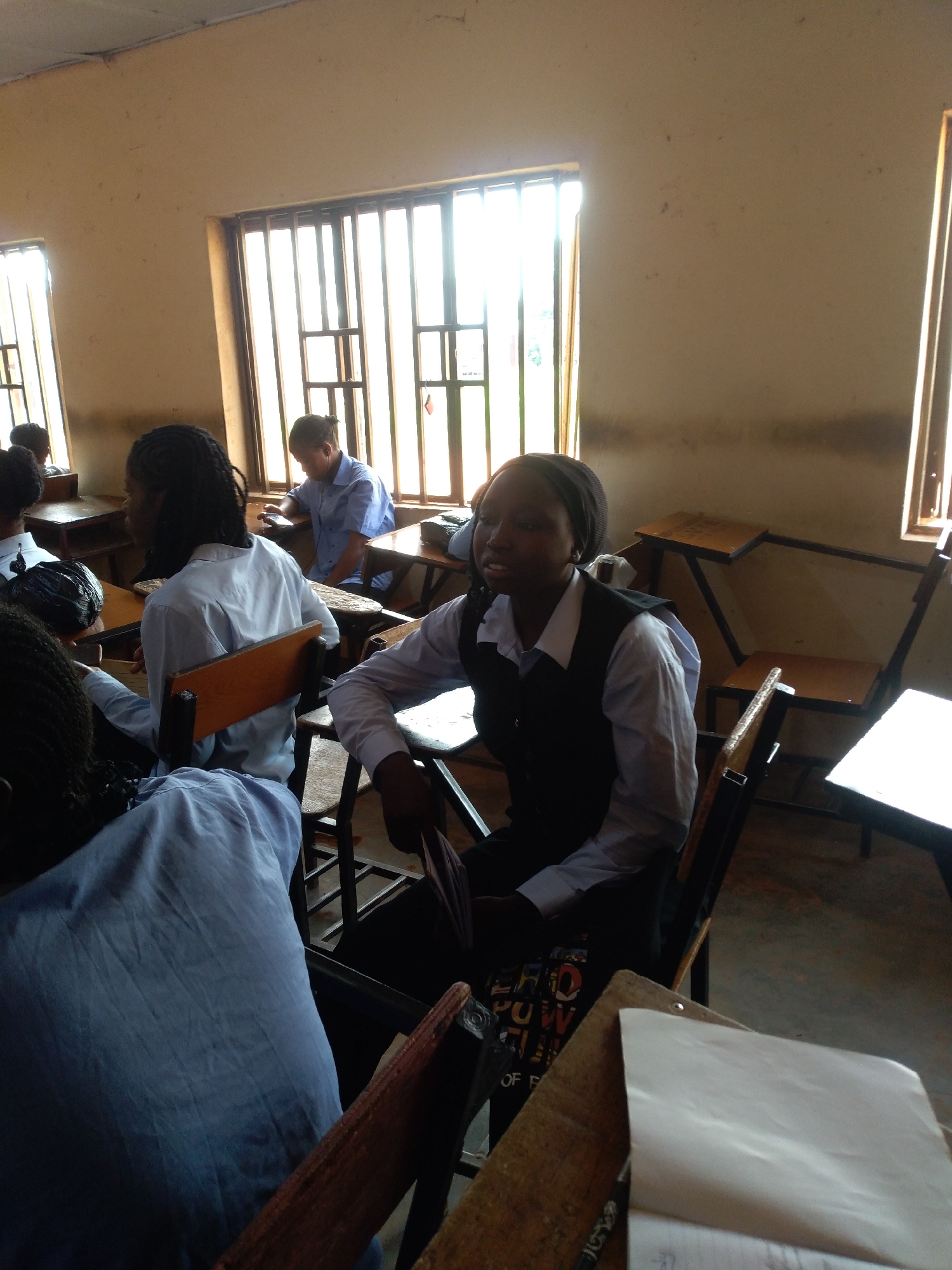 In a classroom in Jos, Nigeria, two young women, likely students, are seated at desks. The woman in the foreground, wearing a light blue shirt and a dark vest, is looking to the right. The woman behind her is wearing a similar light blue shirt. Other students are visible in the background, as well as desks and windows with metal bars. The room appears to be simply furnished and lit by natural light from the windows.