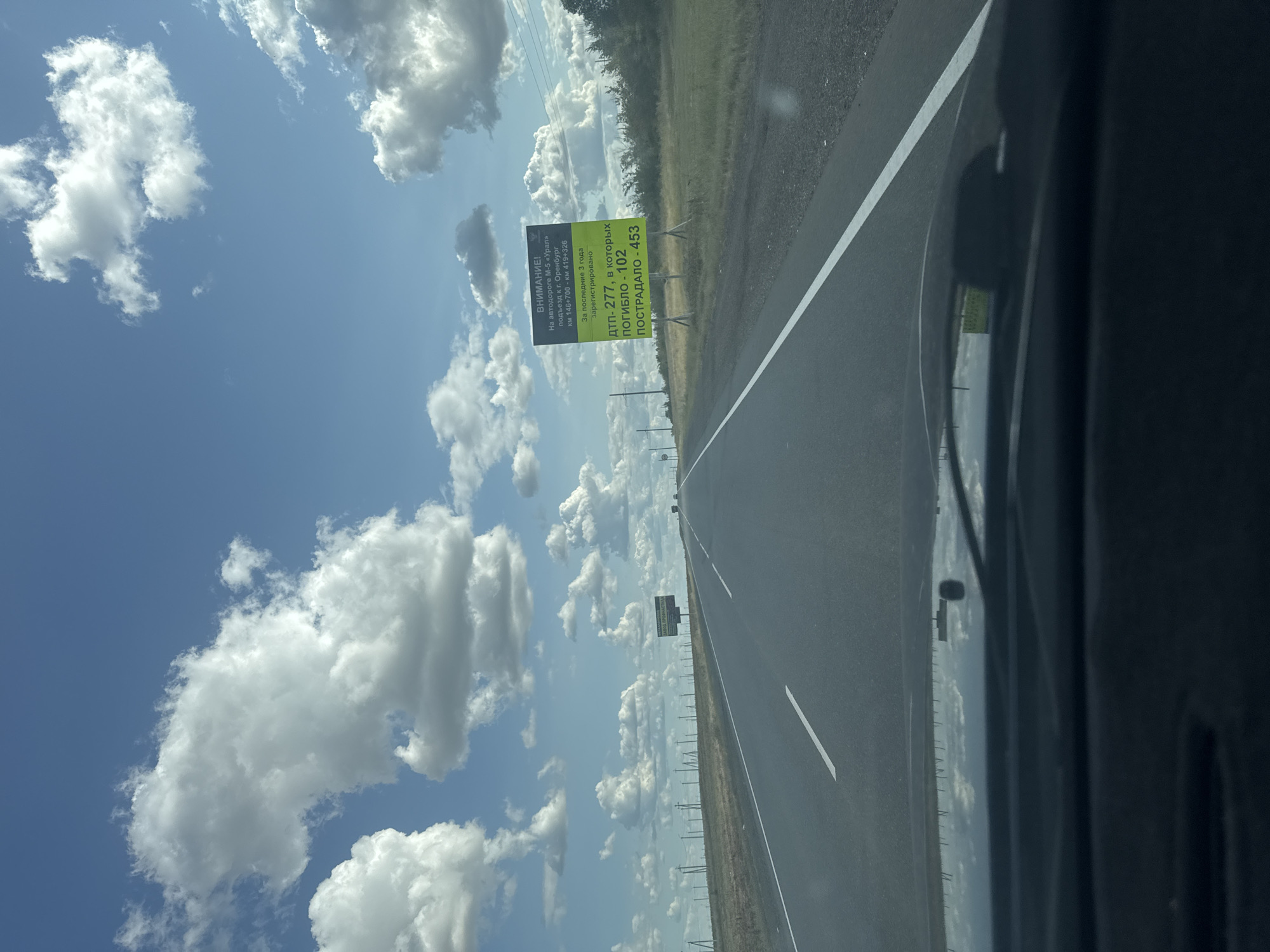 The image shows a road in Novosergiyevka, Russia, on a cloudy day. The road is asphalt and appears to be a highway with multiple lanes. There are power lines on the left side of the road and trees and grass on the right side. A billboard is visible on the right side of the road. The billboard contains text in Russian. The sky is blue with white clouds. The photo is taken from inside a vehicle, as indicated by the dashboard and windshield wipers visible in the foreground.