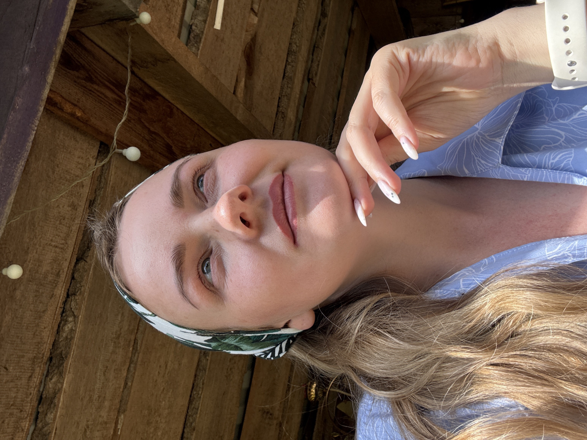 A portrait of a woman with fair skin, light blonde hair, and blue eyes. She is wearing a patterned headband and a light blue floral-print shirt. Her right hand is near her face, with long nails painted with white tips and small decorations. She is positioned against a wooden background, possibly a wall or structure, with string lights visible in the upper left corner. The image is likely taken outdoors in a location in the city of Chaikovsky, Russia based on the user's request.