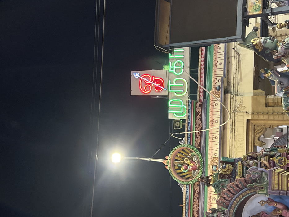 The image captures a nighttime scene outside what appears to be a Hindu temple in Palani, India. The dominant subject is a brightly lit neon sign, composed of a red symbol and green Tamil text. The red symbol depicts a stylized "Vel," the divine spear associated with Lord Murugan. The green neon text reads "முருகர்" (Murugar), the Tamil name for the deity Lord Murugan, for whom Palani is a significant pilgrimage site.
Below and to the right of the sign, intricate temple architecture is visible, characteristic of South Indian gopurams or temple facades. These structures feature multiple tiers, vibrant carvings of deities, and decorative elements in various colors, though some details are obscured by the night and angle. A circular, ornate relief with figures is also visible, likely part of the temple's decorative work.
A bright street light on a pole illuminates the lower left portion of the image, contrasting sharply with the dark night sky. Power lines crisscross the upper part of the frame. There is no specific human activity or event taking place, but the illuminated sign suggests that the establishment, likely a temple or a shop catering to devotees, is open or accessible after dark. The overall impression is one of a bustling religious or commercial area at night.