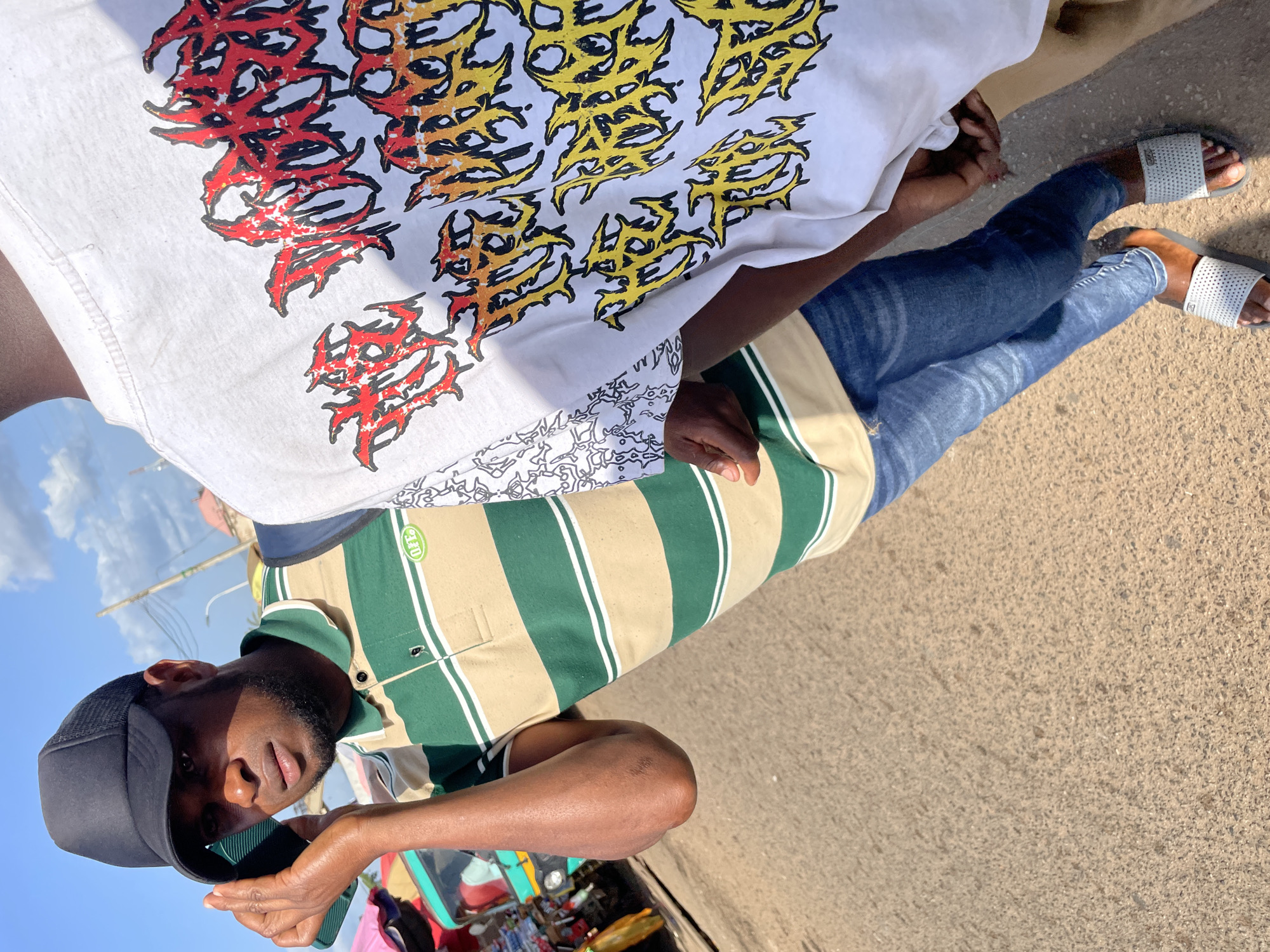Two individuals are captured in a close-up shot in Bukuru, Nigeria. The person on the left wears a black cap, a striped polo shirt in green and tan, and blue denim jeans. They are holding a phone to their ear with their right hand. The person on the right wears a white t-shirt with a graphic print featuring fiery red and yellow lettering. Both are standing on a paved surface, possibly a street or walkway, with a slightly out-of-focus background showing hints of a marketplace with colorful stalls and vehicles. The lighting suggests it is daytime, with shadows indicating a bright sun.