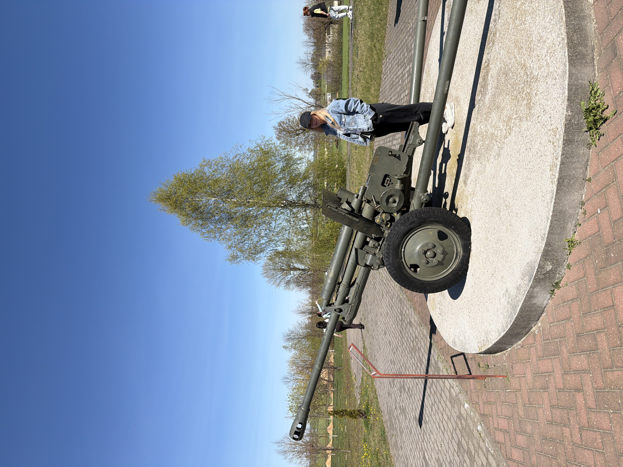 A military gun exhibit rests on a gray concrete slab on a red brick patio in Свіслацкі сельскі Савет, Belarus. A fair-skinned person with long blond hair, wearing a black hat and a denim jacket, leans toward the artillery. To the left is a thin tree with green leaves. Above the exhibit and tree, the sky is clear and blue. In the background, two people stand on a grassy field with several trees. The ground around the brick patio is grassy.