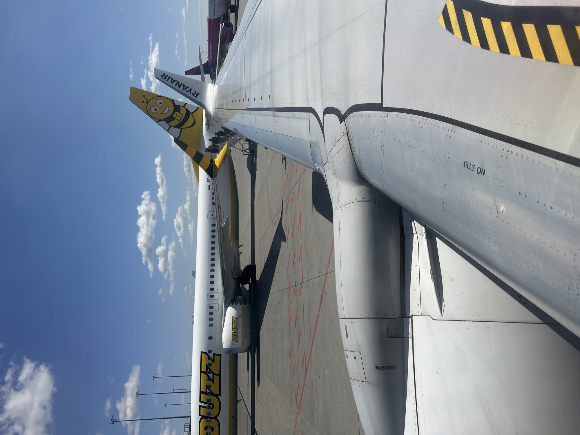 A ground-level, aerial view depicts the wing of a Ryanair airplane from inside another aircraft. The wing is grey with visible rivets and the words "FLIS ON" are printed in black letters. Next to the Ryanair plane is a plane painted with the word "BUZZ" in large yellow letters, and a cartoon bee is on the tail of the plane. A third plane, with magenta coloring, is partially visible. The sky is blue with white clouds. The location is Wrocław, Poland.