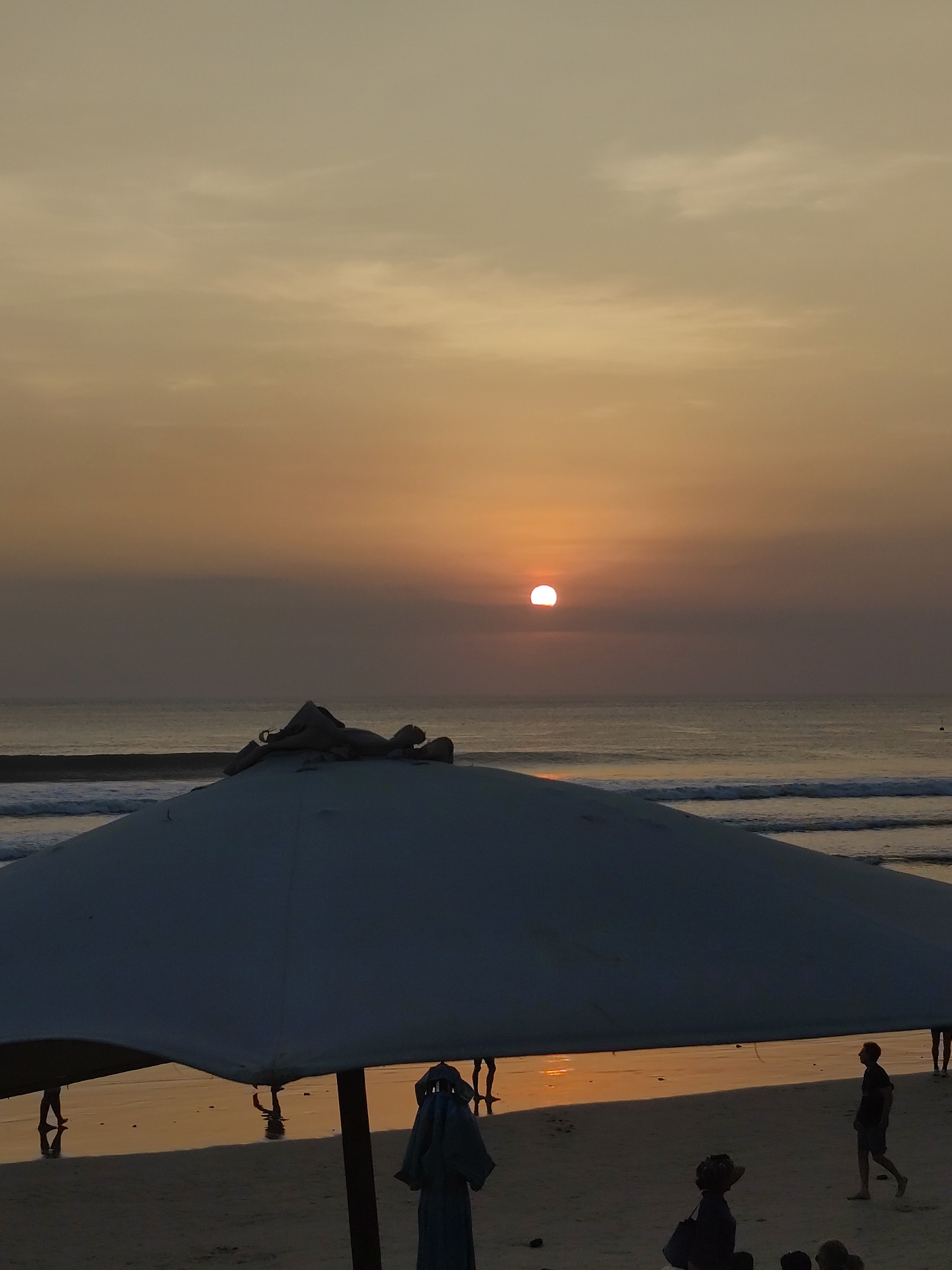 The image captures a sunset scene at an unknown beach location. The sun, a bright orb, is positioned low in the sky, casting a warm orange glow that reflects on the calm ocean and wet sand. The foreground is dominated by a large, dark umbrella, its underside illuminated by the sunset. Several people are visible on the beach; some are walking, while others are silhouetted against the sand and water. In the distance, waves gently roll onto the shore. The overall atmosphere is serene and tranquil.