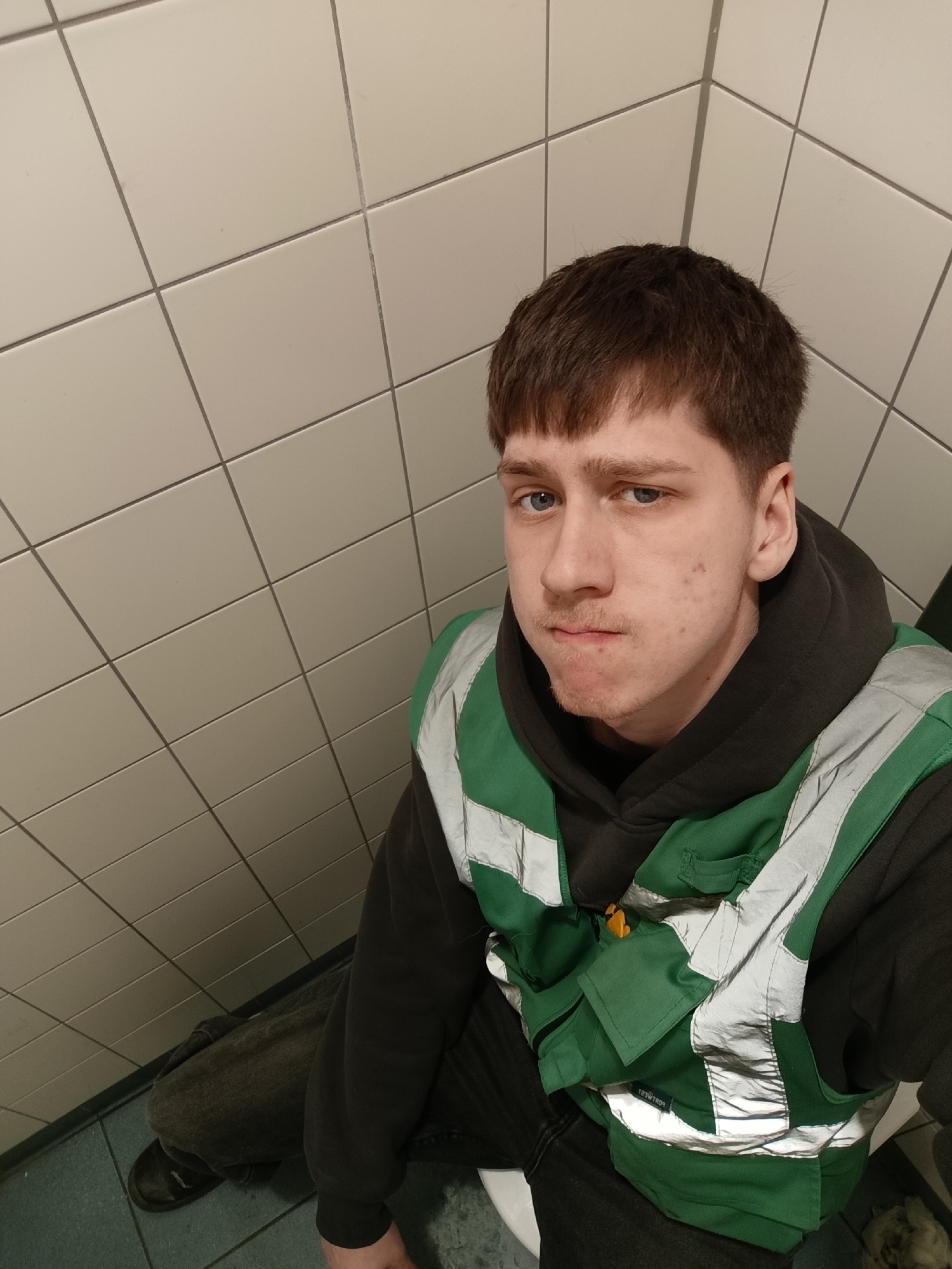 A young Caucasian male with short brown hair, light blue eyes, and a sparse mustache is seated in what appears to be a restroom in Duiven, Netherlands. He is wearing a dark-colored hoodie under a green safety vest with reflective stripes. The restroom has white tiled walls and blue-green tiled flooring. His legs are positioned toward the bottom left corner of the frame, suggesting a seated posture. The camera is positioned above him, angled downward.