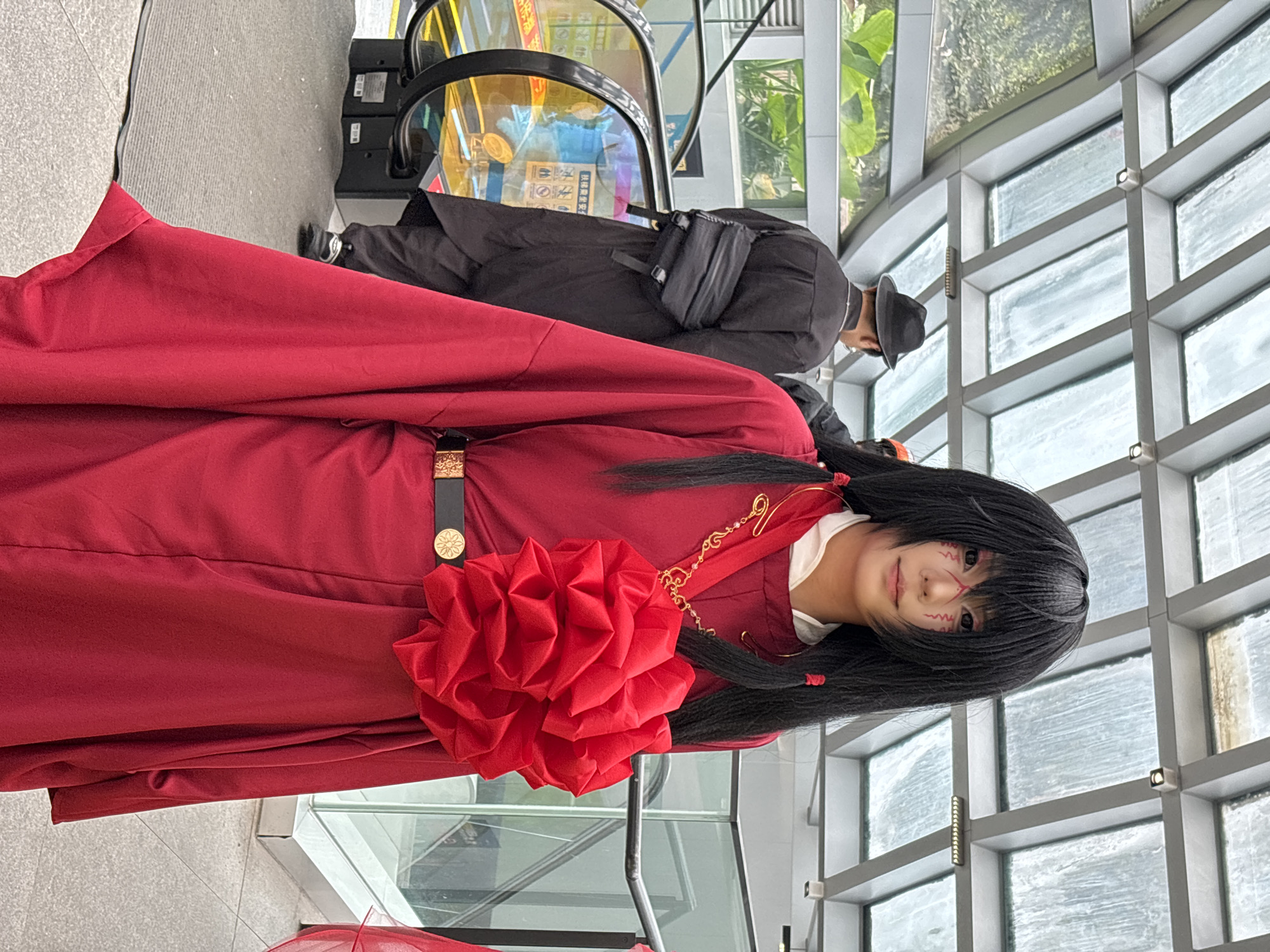 A person in a red costume with a large red flower pinned to it stands in front of a wall of windows. The person has black hair and facial markings. Behind them is an escalator and another person in black clothing and a hat. The location is possibly in the city of Liangjiang, China.