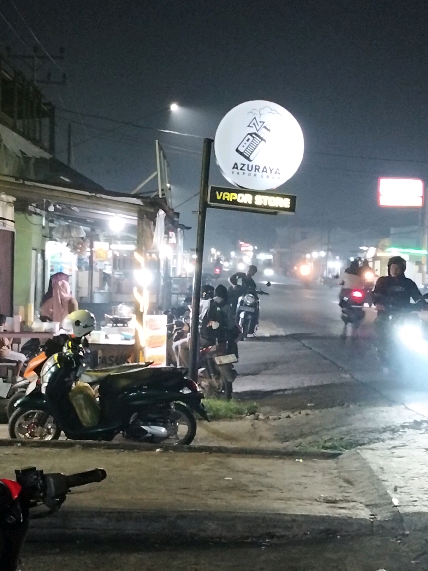 The image depicts a street scene at night in Arang Limbung, Indonesia. A prominent circular white sign with a black logo and text reading "AZURAYA VAPOR STORE" is mounted on a pole. Below it, a horizontal yellow sign with illuminated text states "VAPOR STORE." Several motorcycles are parked and in motion. In the foreground, a dark-colored scooter with a yellow bag on the seat is parked. A red motorcycle handlebar is visible in the lower left corner. In the background, individuals are seen on motorcycles, some with illuminated headlights. Buildings line the street, with lights indicating a commercial area, possibly featuring food stalls. The overall atmosphere is dimly lit, characteristic of a street at night, with artificial lighting from signs and vehicles dominating the scene.