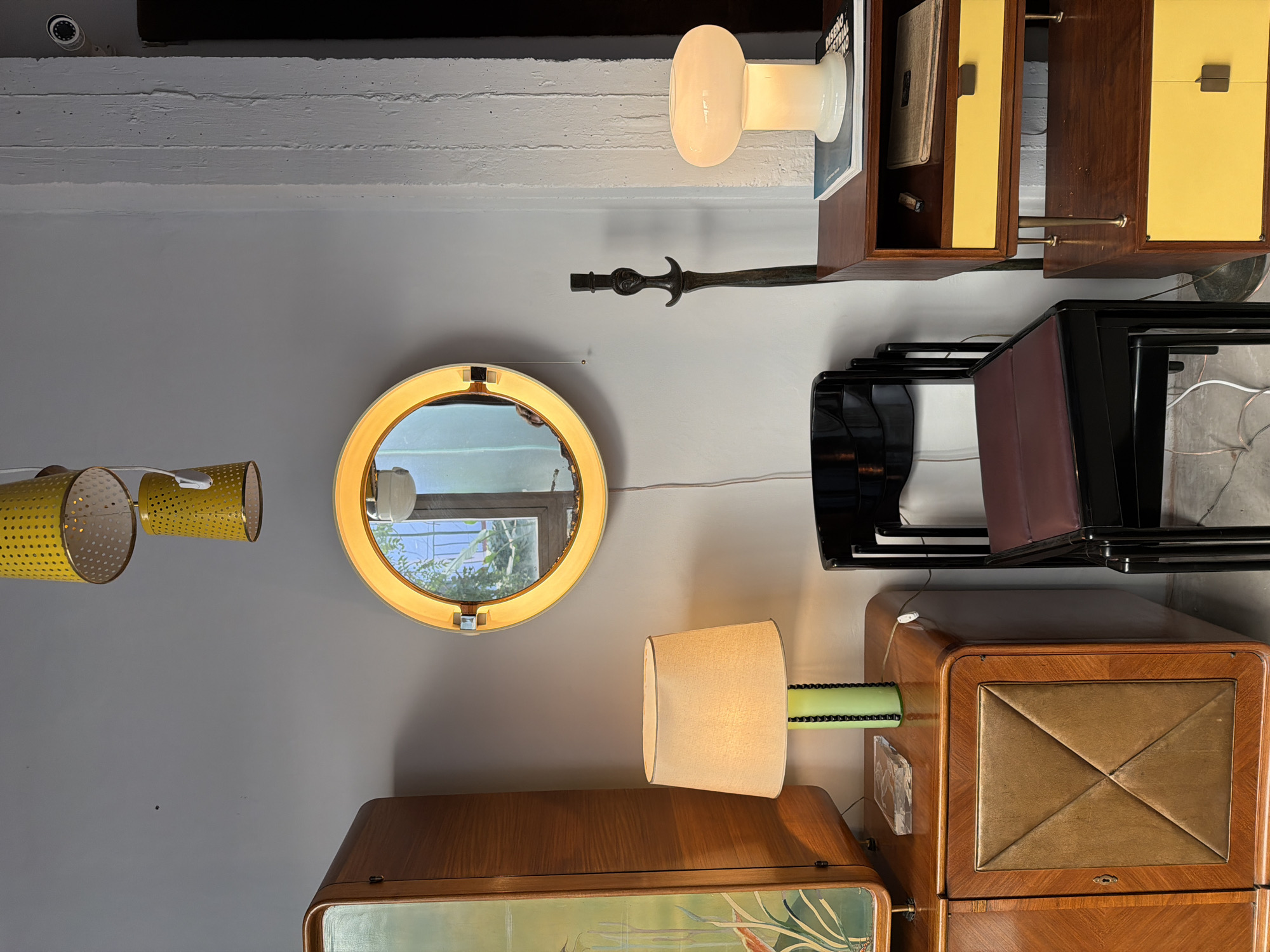 This media file captures an interior scene, likely a vintage furniture and lighting showroom or shop in Buenos Aires, Argentina. The room features light grey walls and a white concrete ceiling with visible beams.
Prominently displayed are various pieces of furniture and lighting fixtures, suggesting a mid-century modern or Art Deco aesthetic. On the upper right, a wooden wall-mounted cabinet with two yellow-fronted compartments holds books, one of which has "DISEÑO INDUSTRIAL" (Industrial Design) visible on its spine. A glowing white mushroom-shaped lamp is mounted above this cabinet. Below it, a glossy black, possibly lacquered, table or cabinet with dark reddish-brown accents is visible.
A decorative, dark-colored sword-like object with an ornate handle is mounted horizontally on the wall. Central to the image is a large, round mirror with a wide, golden frame, reflecting an outdoor scene with greenery and a window. To the left of the mirror, two yellow, perforated cylindrical pendant lamps hang from the ceiling.
Towards the bottom right, several wooden cabinets with rounded edges are arranged on the floor. One cabinet features a distinctive gold-colored, padded X-shaped design on its front panel. Another has a table lamp with a light green, columnar base and a simple beige drum shade. Wiring for some of the lamps is visible. In the upper right corner of the ceiling, a black security camera is mounted.
The overall impression is a curated display of design objects, without any human presence or specific activity taking place. The lighting is artificial, contributing to the indoor display setting.