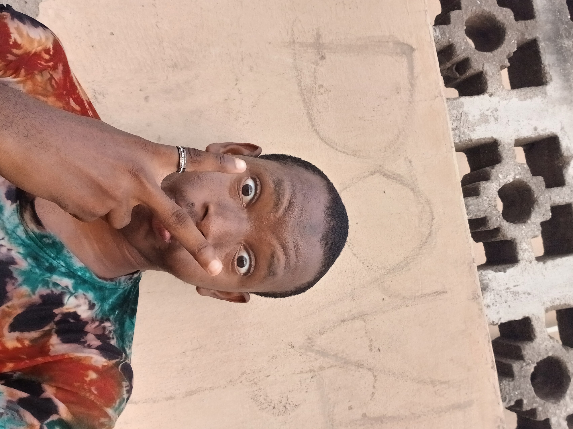 A man stands facing the camera. He is holding his fingers in front of his eye. He is wearing a ring. The man is wearing a tie-dye shirt. The background is an unpainted wall and a concrete structure with hollowed-out sections. This scene appears to be in Lagos, Nigeria.