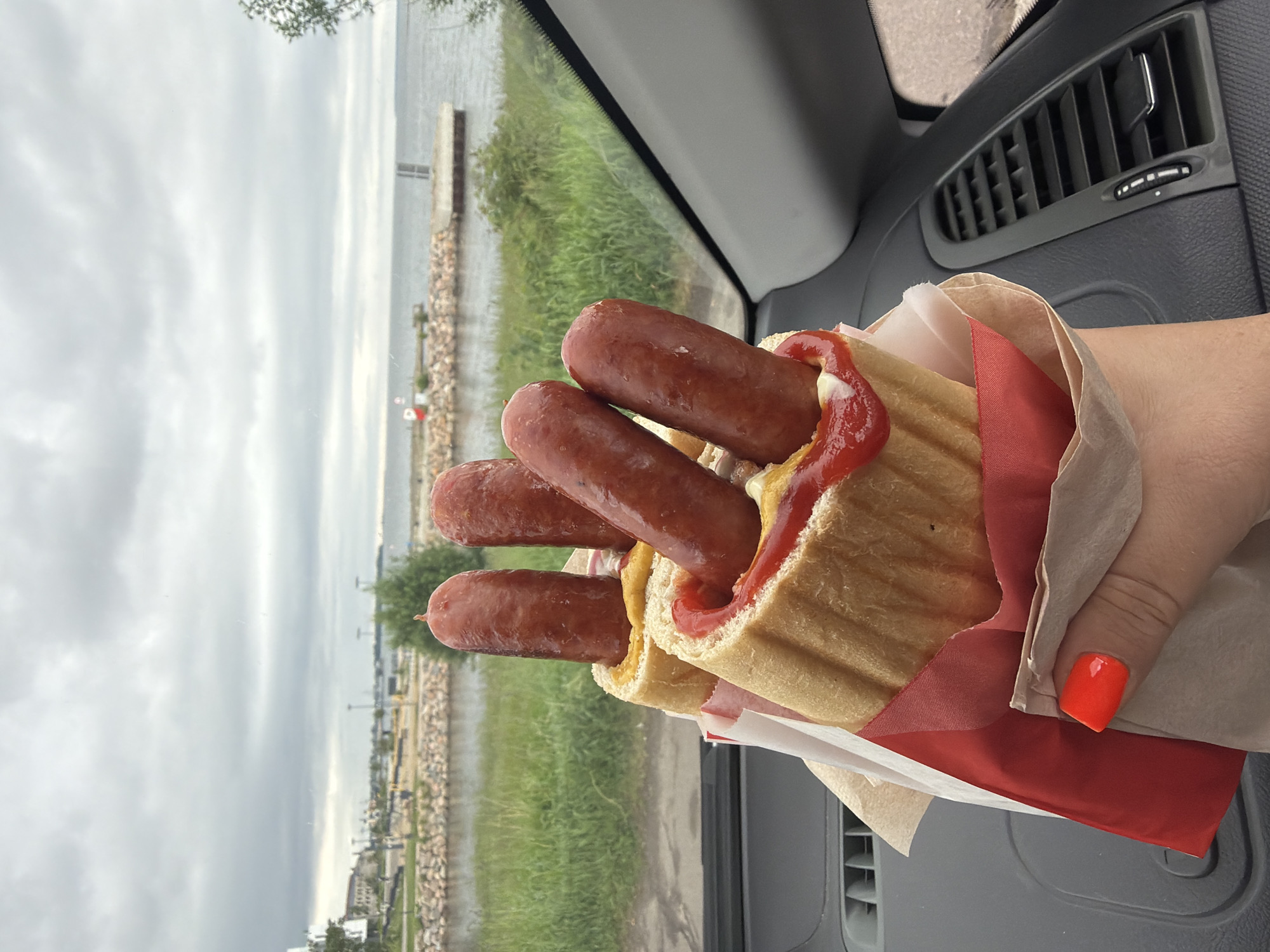 A close-up shot shows a person's hand with bright orange fingernails holding a large hot dog with multiple sausages, ketchup, and mustard. The hot dog is wrapped in paper and appears to be made with a long, split bun. In the background, a body of water, possibly the Baltic Sea, is visible under a cloudy sky. Along the shoreline, there's a rocky embankment, green grass, and a distant pier or breakwater with a white and red marker. The scene is framed by the interior of a vehicle, with a dashboard vent visible on the right. The location is the Pirita beach area in Tallinn, Estonia, a popular spot for locals and tourists.