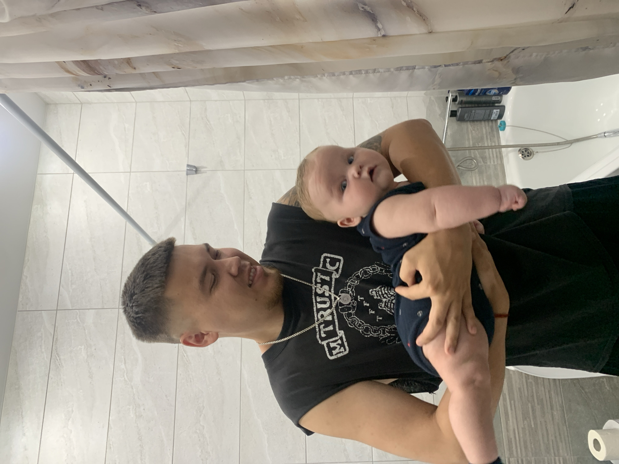 In a tiled bathroom, a man with short, buzzed hair and a black sleeveless t-shirt holds a baby. The baby is wearing a dark blue outfit. The man is smiling as he looks down at the baby, who is looking towards the camera with wide eyes. A shower curtain hangs to the right of the frame, and a bathtub is visible in the background. A roll of toilet paper sits on a surface in the lower left corner. The overall impression is of a domestic scene, likely in a residential building within the city of Stavropol, Russia, given the prompt's context.