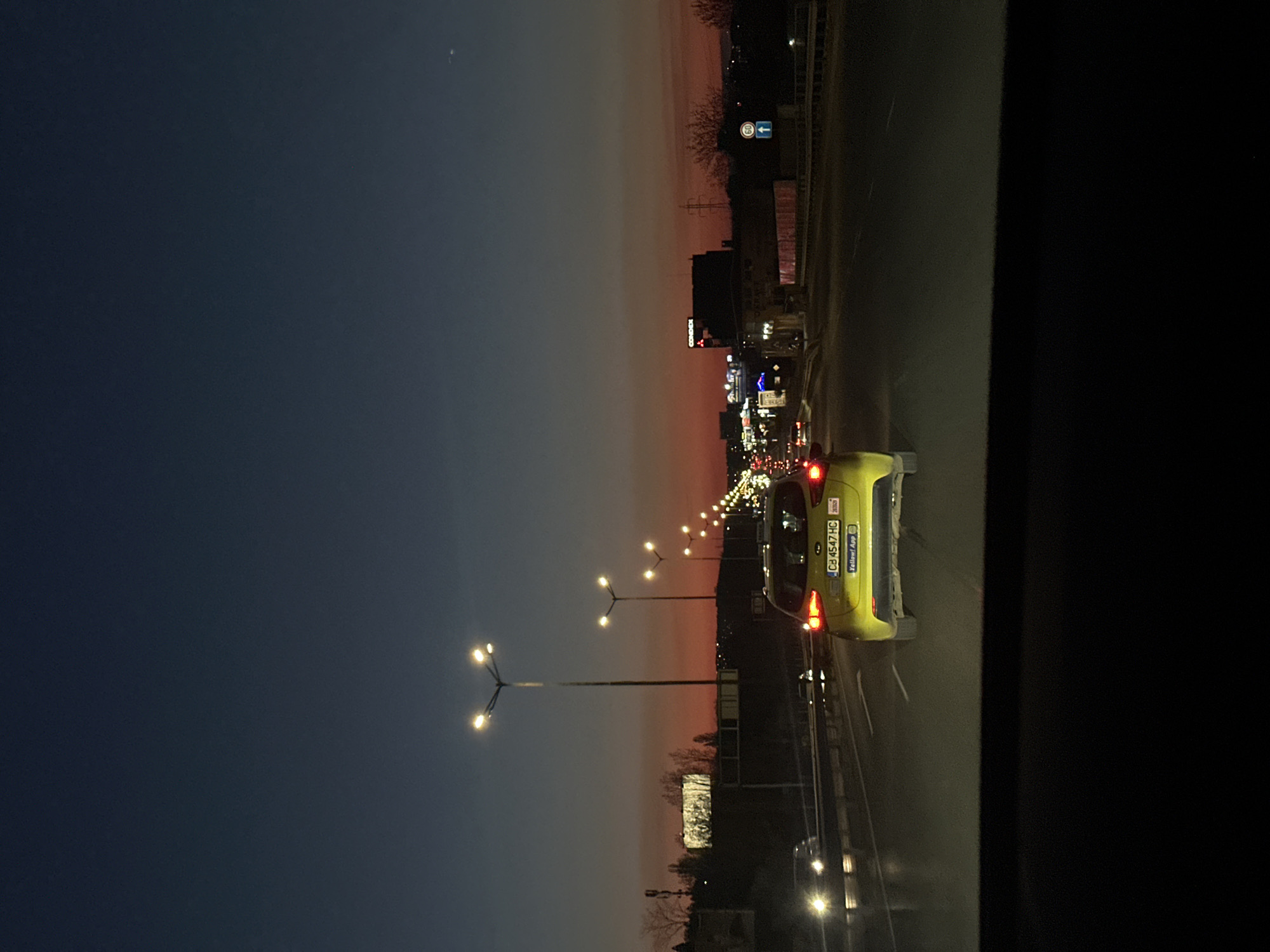 This image captures an urban road scene at dusk, viewed from inside a vehicle, looking forward. The sky transitions from a deep dark blue at the top to a warm orange and pink near the horizon, suggesting either sunset or sunrise.
In the foreground, a bright yellow car, likely a taxi or ride-share vehicle, is prominently visible. It displays a Bulgarian license plate "CB 4547 HC" and text on its rear bumper that reads "Yellow! App" and "20202". Its red taillights are illuminated.
Along the roadside, several distinctive streetlights feature a three-light design, casting bright white light. Further in the distance, a collection of buildings is visible, some with illuminated signs, one of which appears to say "CARGOS". A blue road sign with a white arrow and the number "80" is visible on the right, indicating a speed limit or direction. A metal guardrail runs parallel to the road. The scene suggests movement on a multi-lane road or highway in an urban setting as artificial lights begin to dominate the environment. No people are discernible in the image. The license plate confirms the location context of Sofia, Bulgaria.